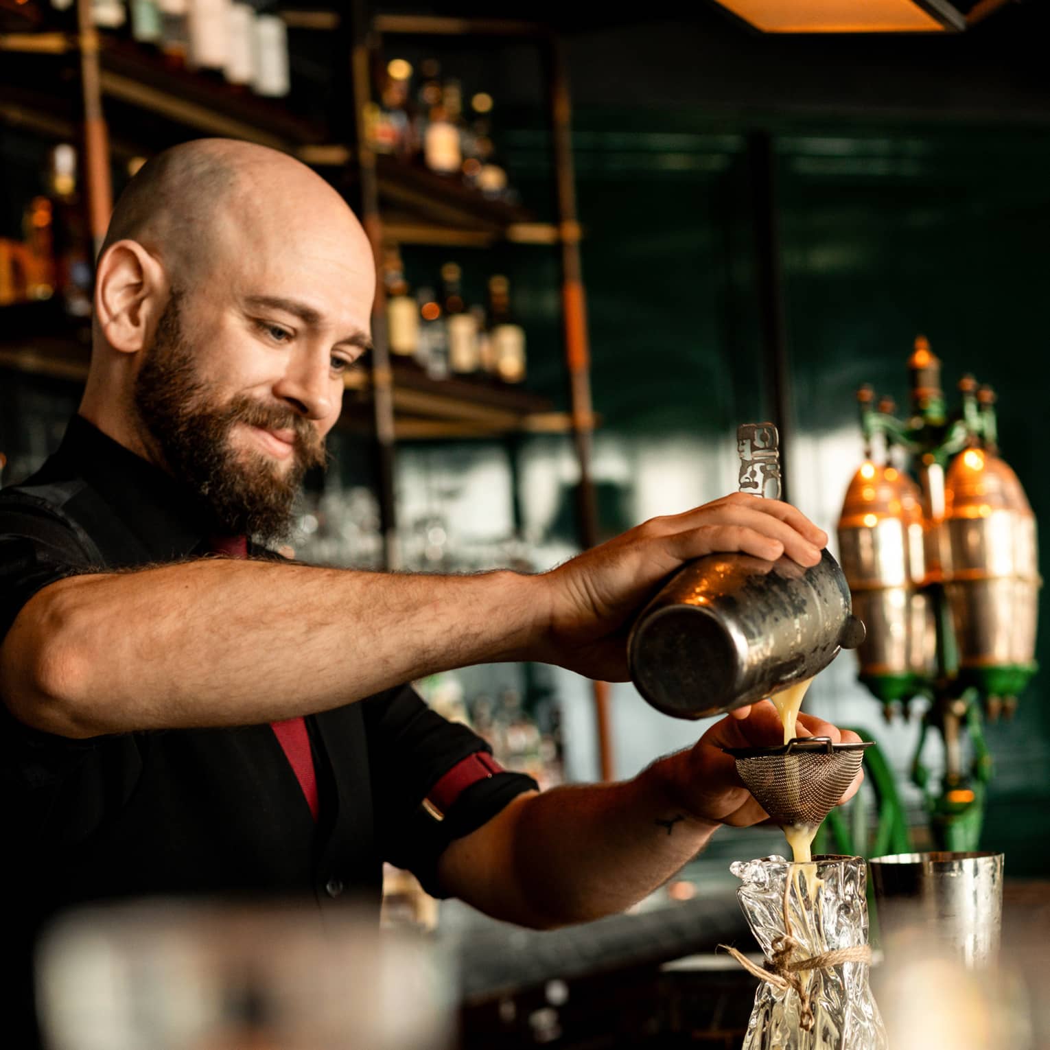 A smiling bartender pours a milky liquid through a strainer into a sculpted blown-glass tumbler, in a warm bar setting.