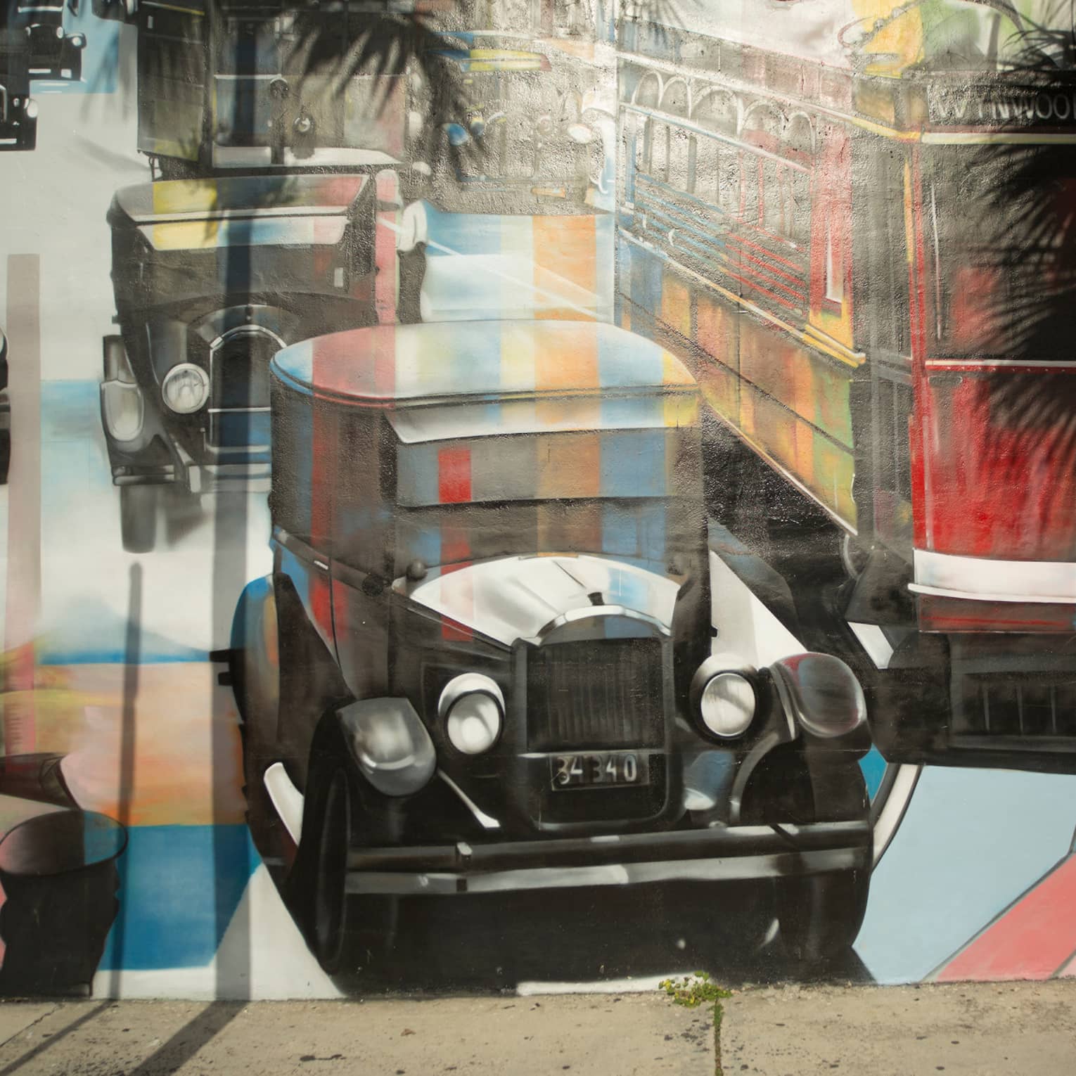 Palm trees cast shadows on a mural of vintage cars and a bus painted in a kaleiscopic pattern of colours and grey shading.