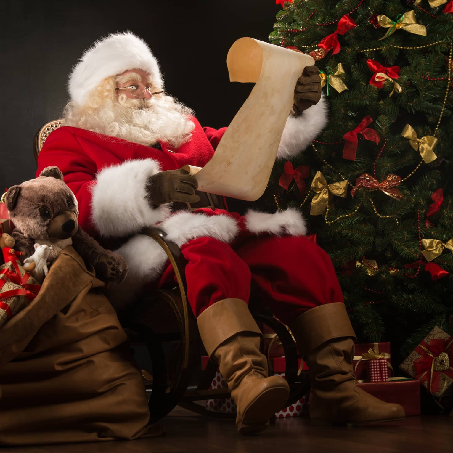 Santa Claus seated in large rocker holding a scroll, flanked by decorated Christmas tree with gifts and overflowing gift bag