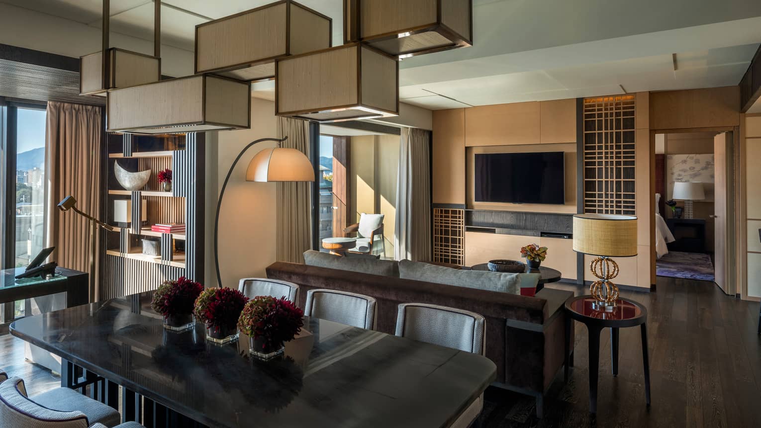 Residential suite with dining table, sofa, wall-mounted TV, floor lamp, bookshelf, desk with leather chair and floor-to-ceiling views of the city skyline