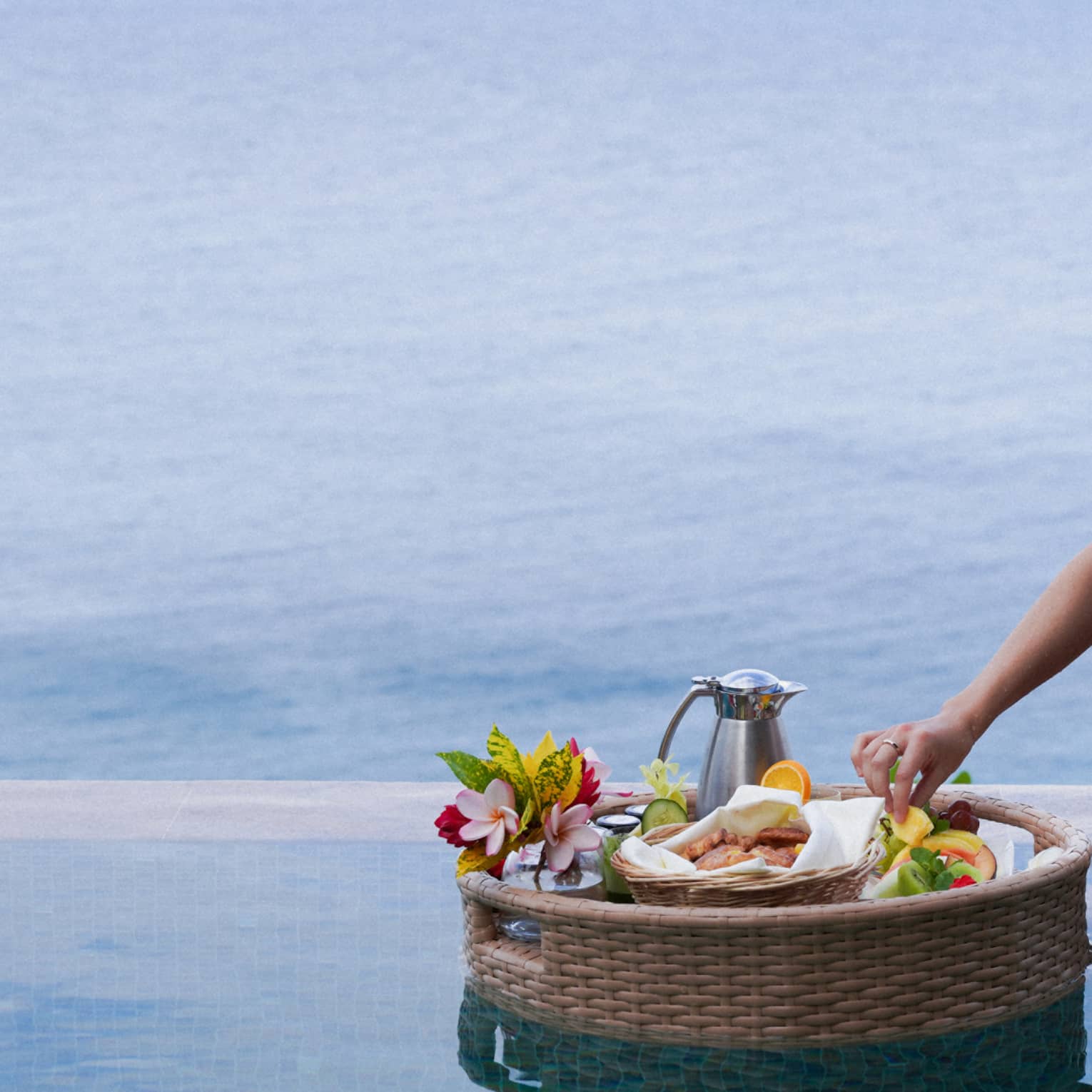A hand delicately reaches into a floating basket of food, fruits, flowers and a carafe in a pristine blue ocean-view pool.
