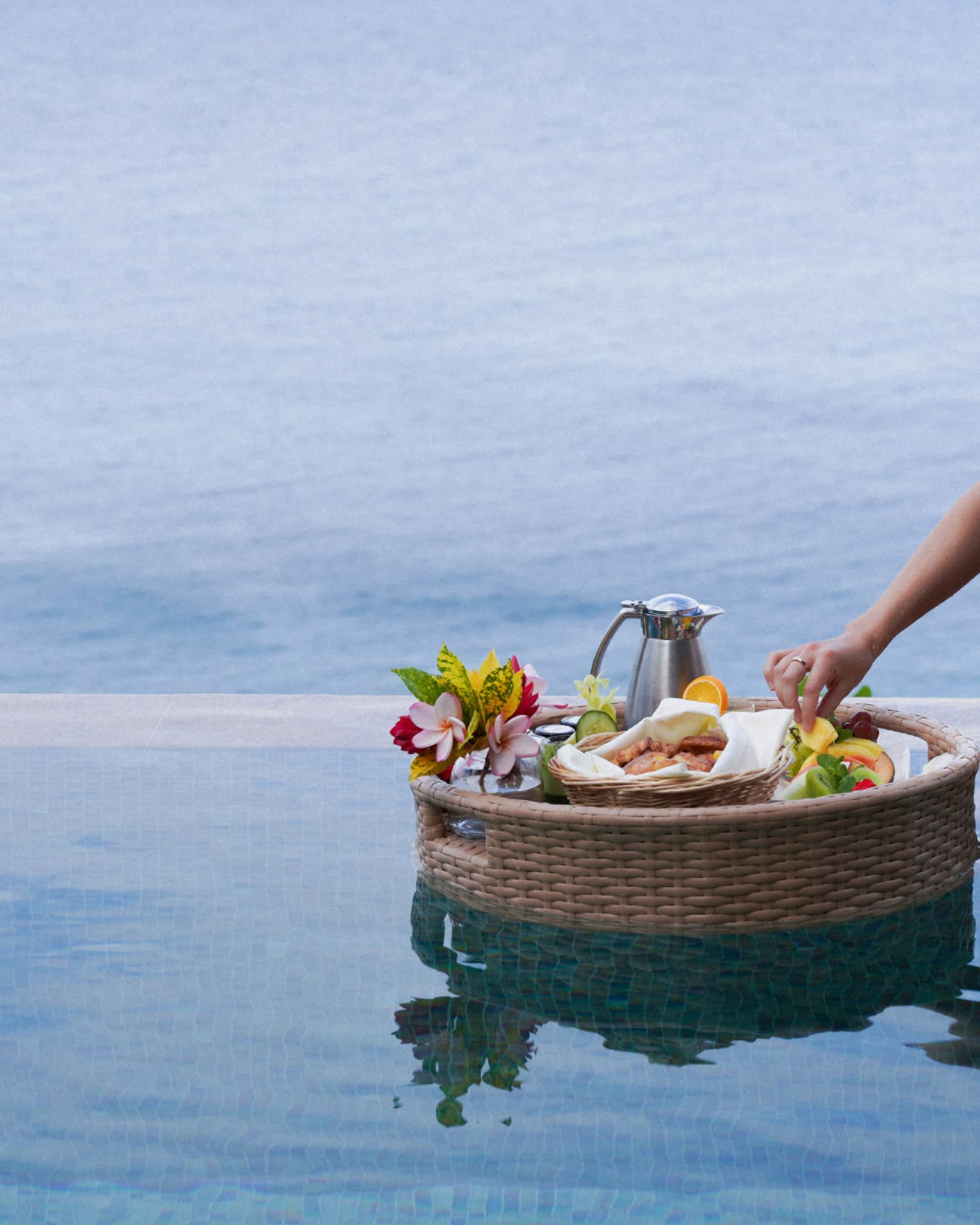 A hand delicately reaches into a floating basket of food, fruits, flowers and a carafe in a pristine blue ocean-view pool.