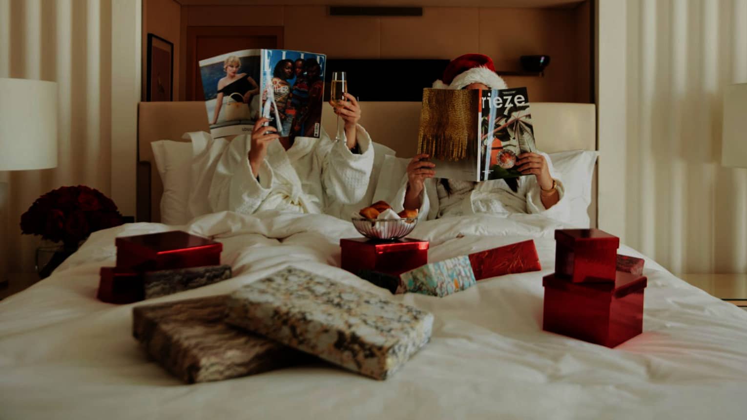 Couple in white bed covered in Christmas gifts, reading magazines