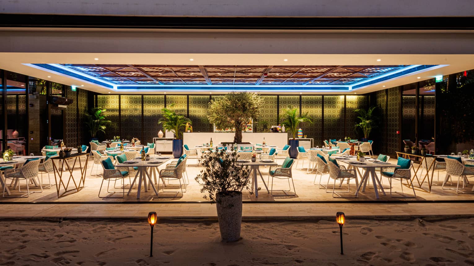 Nighttime view of restaurant dining room open to the beach on one side and surrounded by glass windows on the other sides with a tray ceiling above featuring an inset blue light