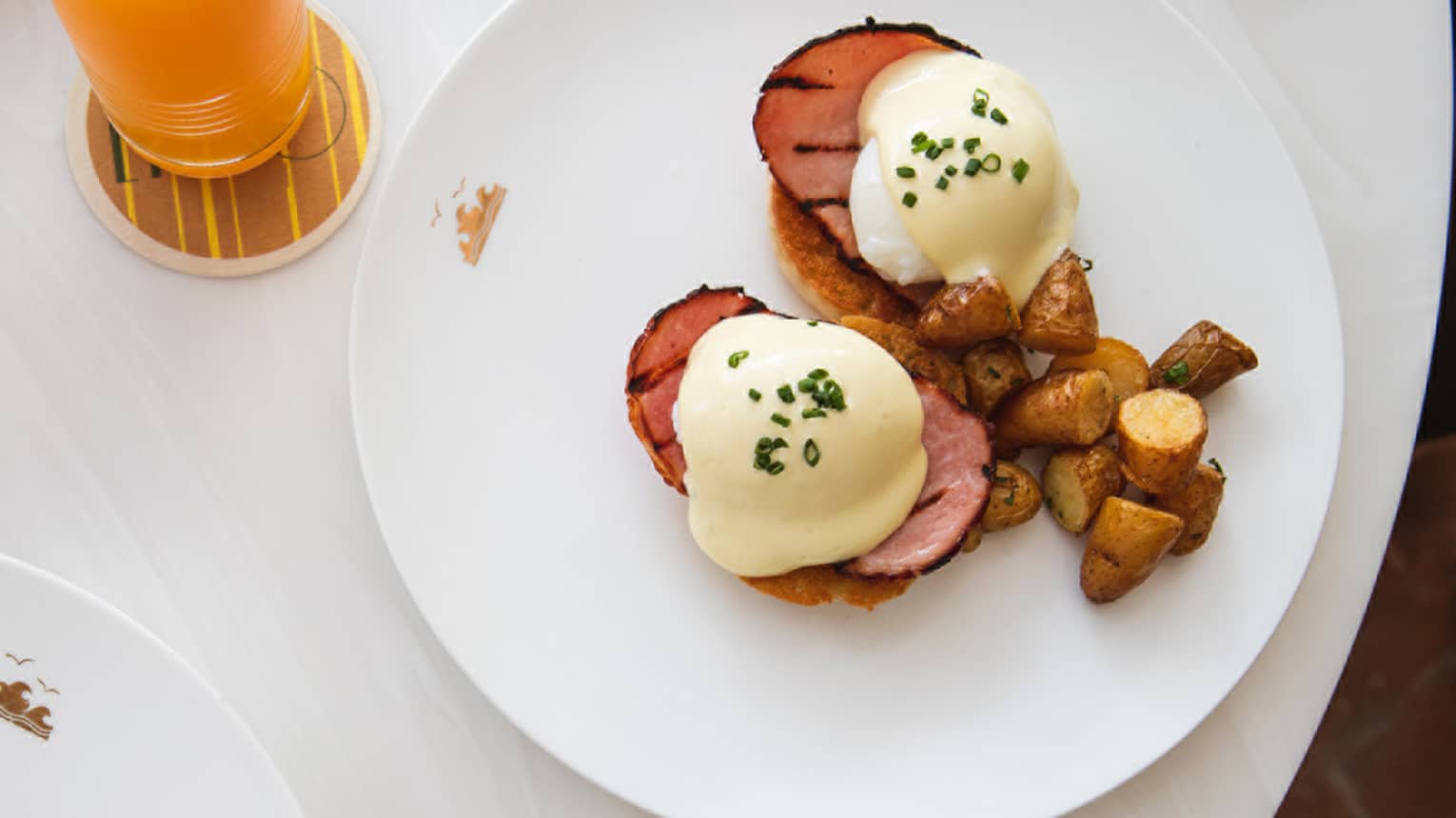 Two plates, one with eggs Benedict and a side of roasted potatoes and one with chia seed pudding topped with berries, sit on a white marble table next to an orange flower in a vase