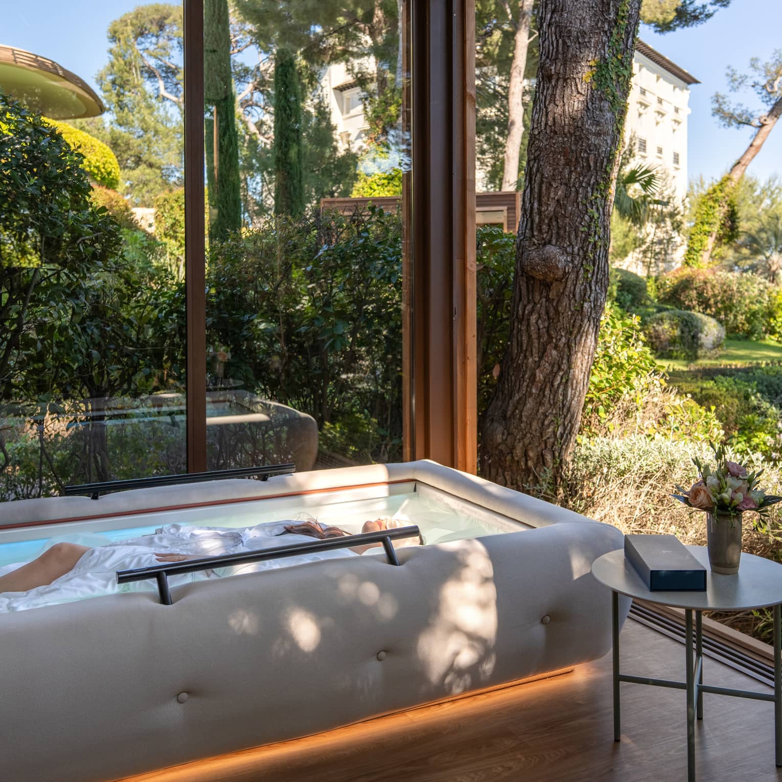 A serene glass-enclosed spa space with a small, raised, oblong pool. Trees and greenery are visible through the glass walls and surround the space.