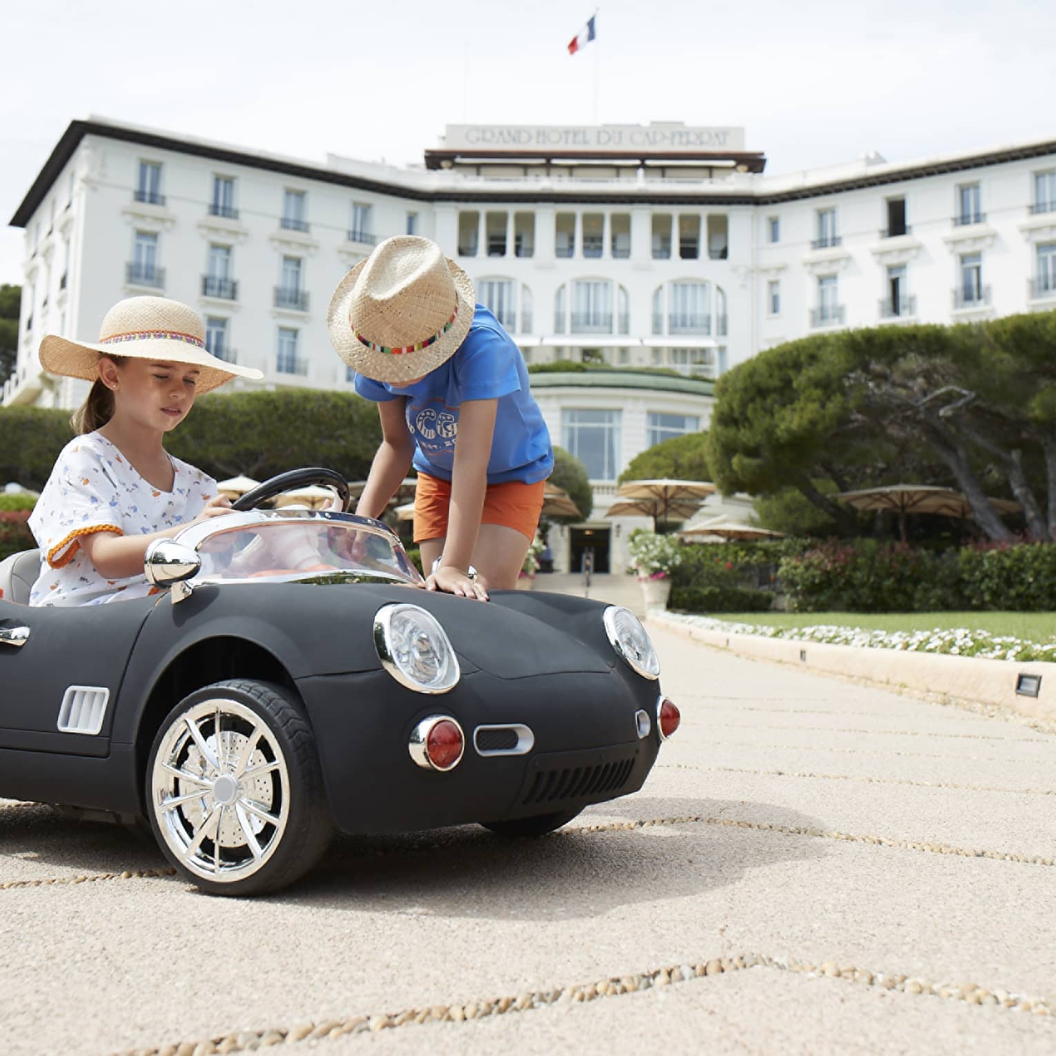 Girl wearing floral dress and straw hat driving black luxury mini convertible, boy in straw hat leaning over, hotel in backdrop