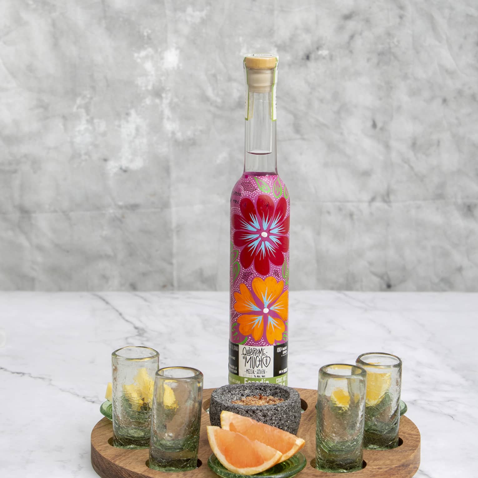 Brightly floral-painted mezcal bottle on wooden tray with four shotglasses, stone spice bowl and lemon and grapefruit wedges.