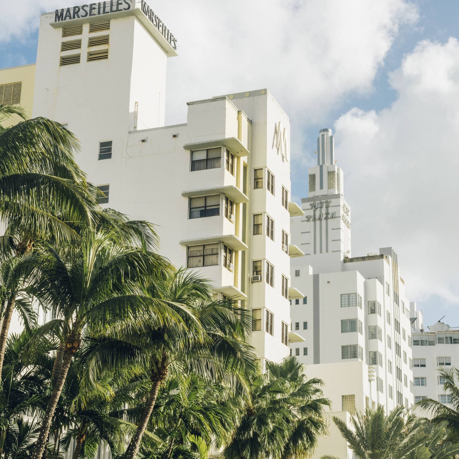 Art Deco buildings with palm trees in front under a bright, partly cloudy sky.