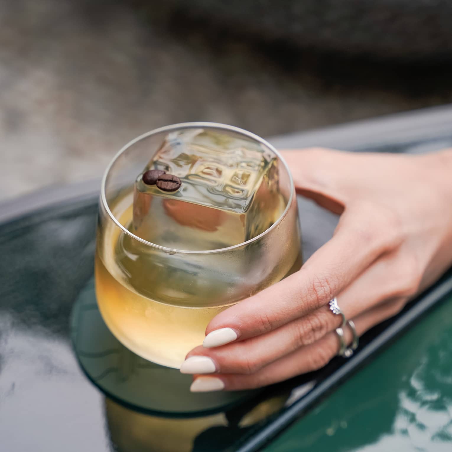 An outstretched hand holds a tan-hued cocktail in a rounded rocks glass