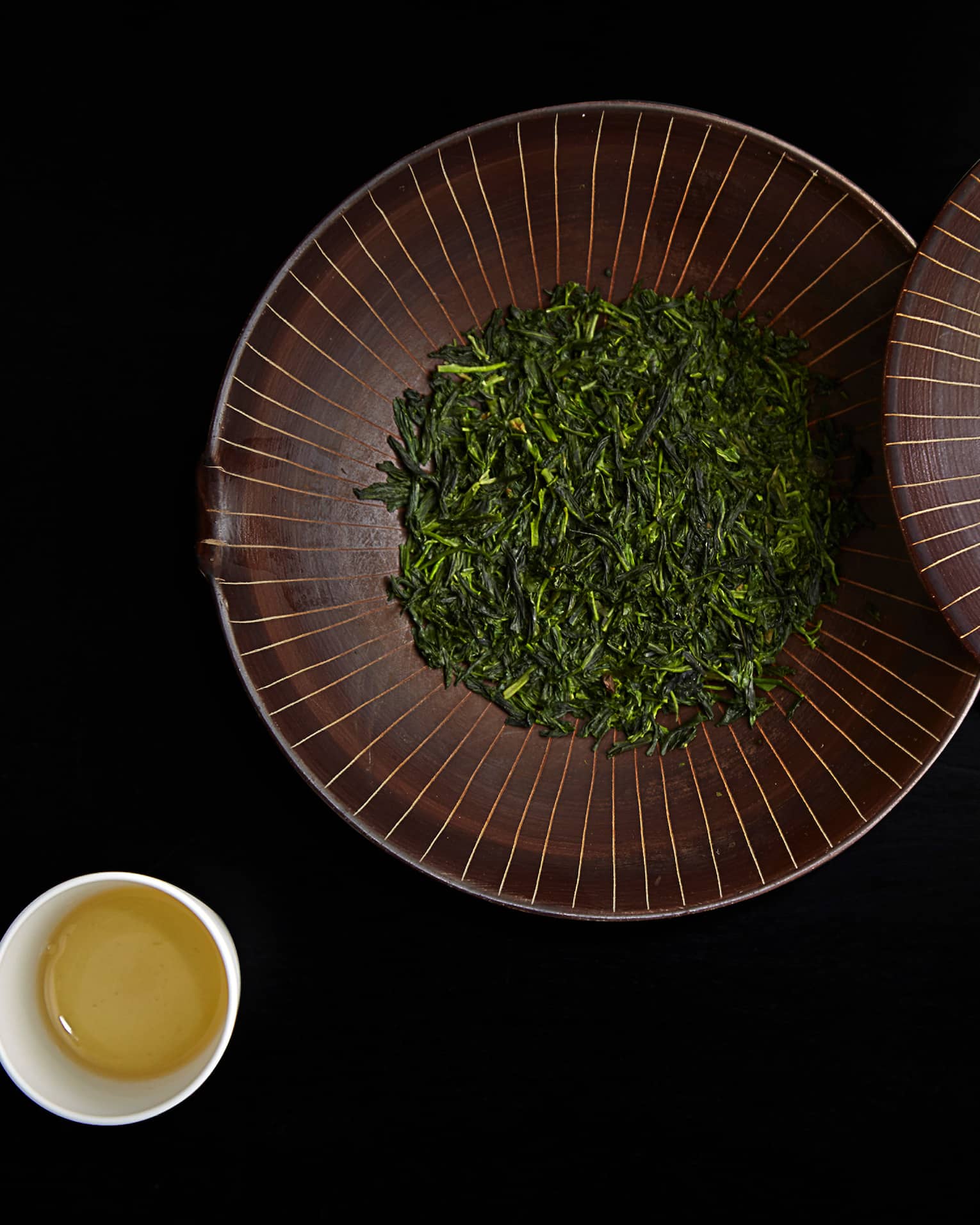 Dried green leaves in large bowl with lid by small cup of tea