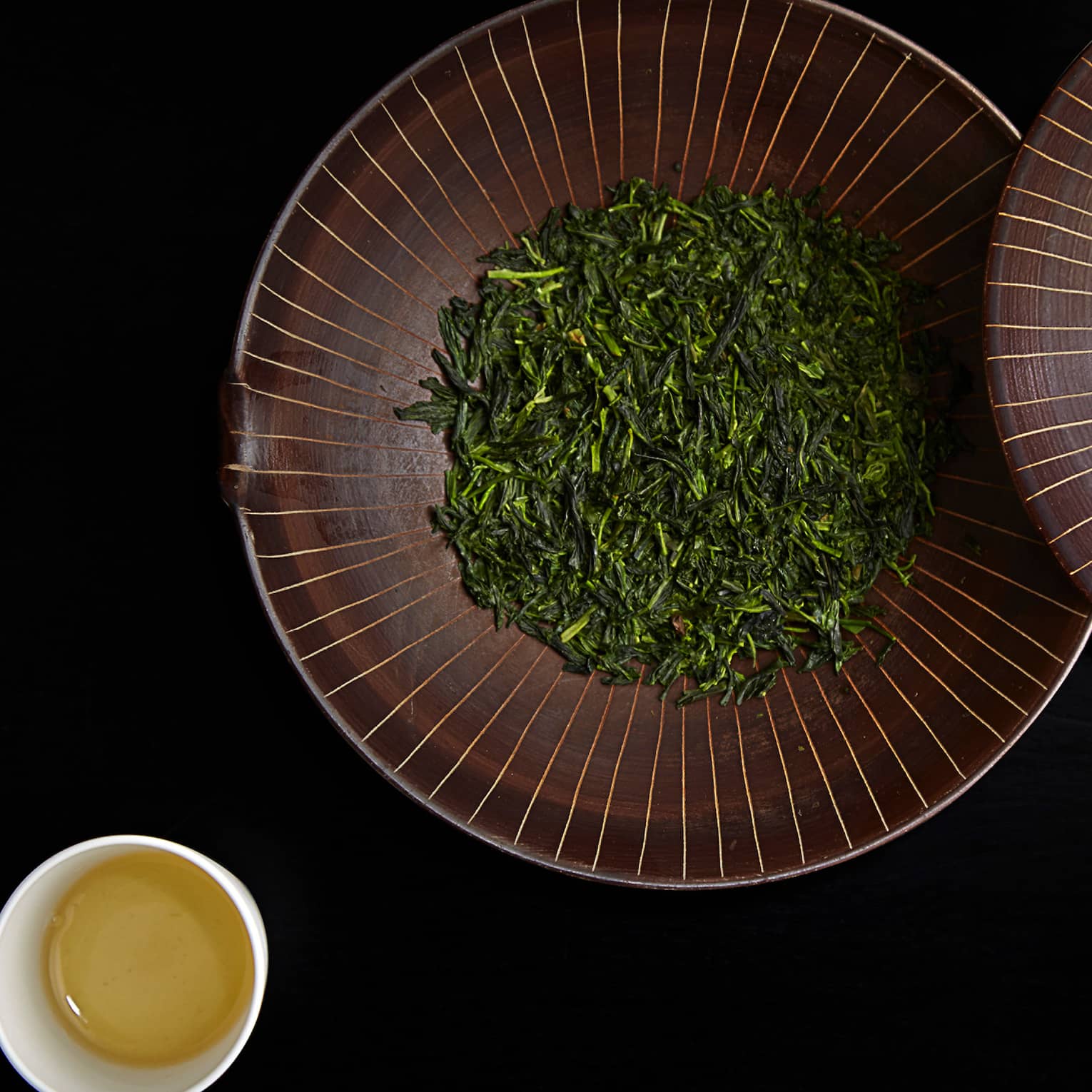 Dried green leaves in large bowl with lid by small cup of tea