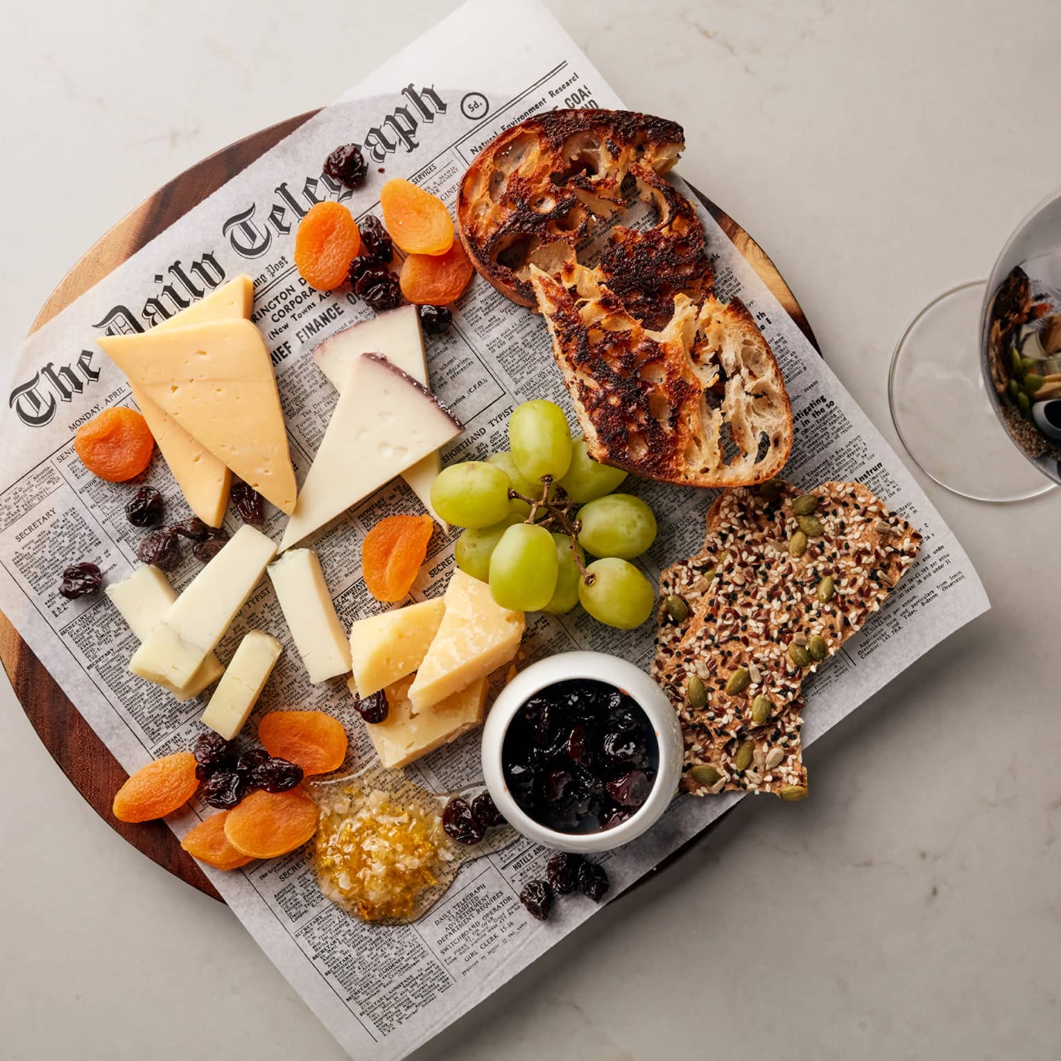 Cheese, grapes, crackers and bread on newspaper on platter next to glass of red wine