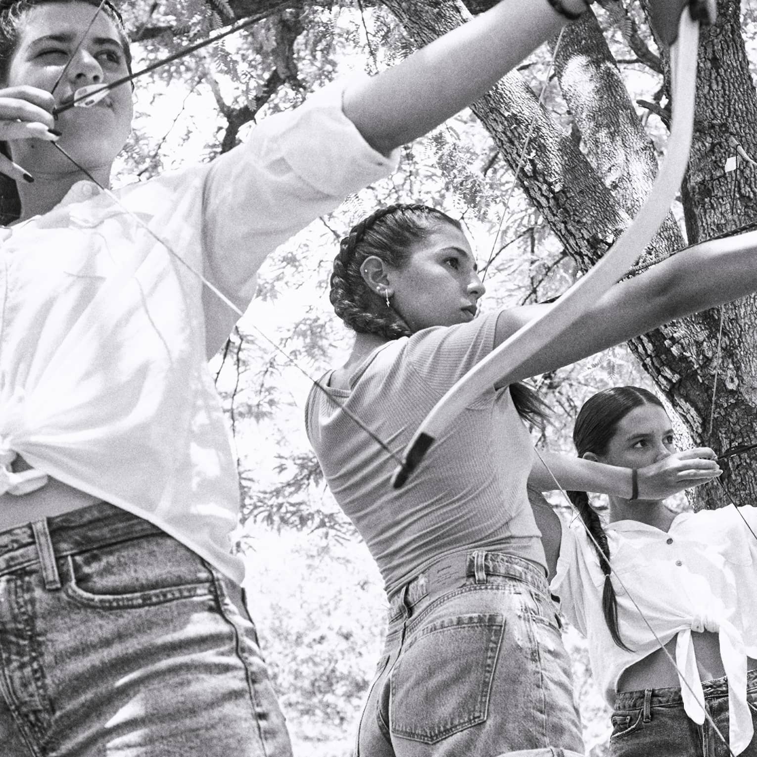 Black and white photo of three young girls aiming traditional archery bows in a forest