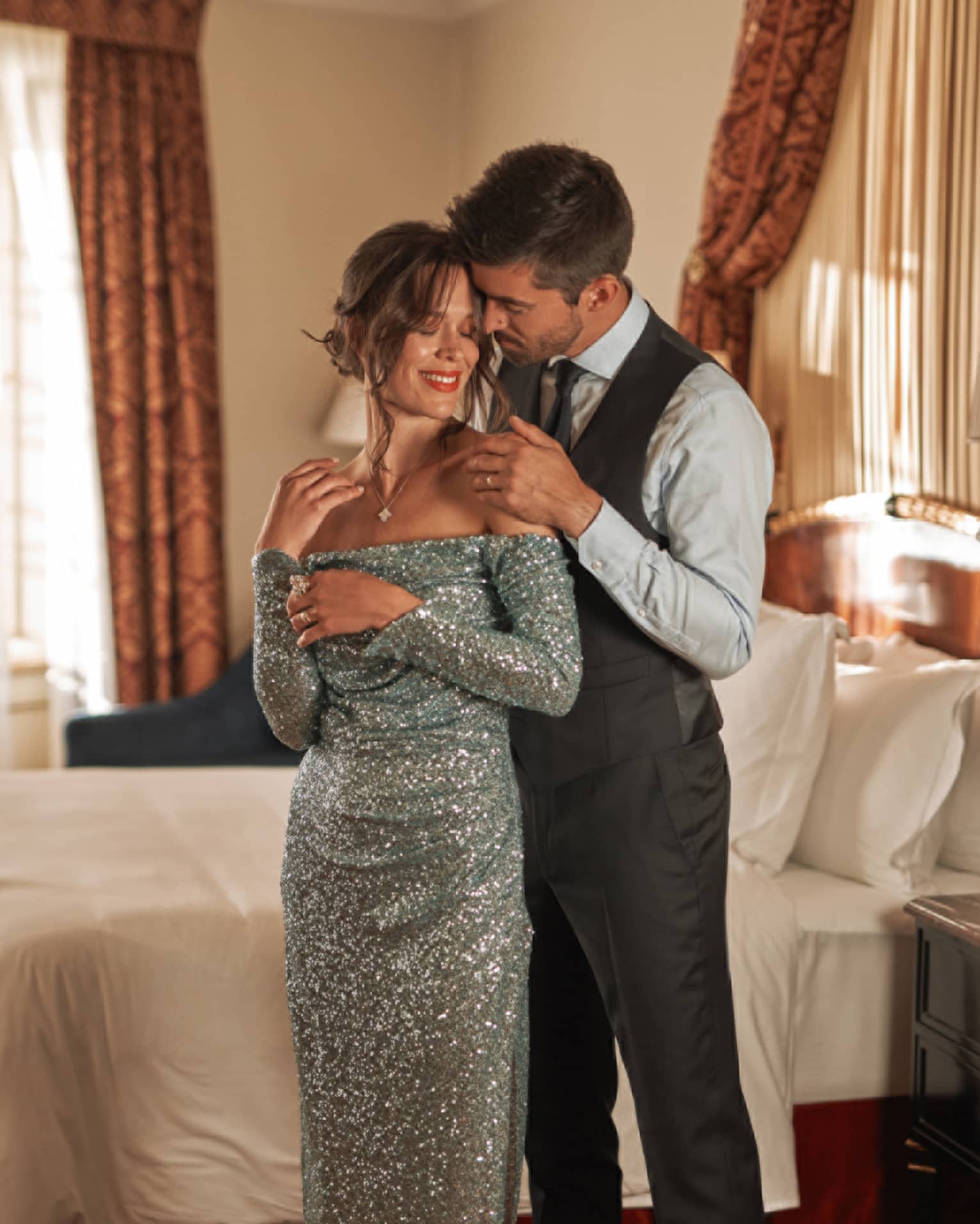 Couple in black tie attire embrace in front of bed in guest room