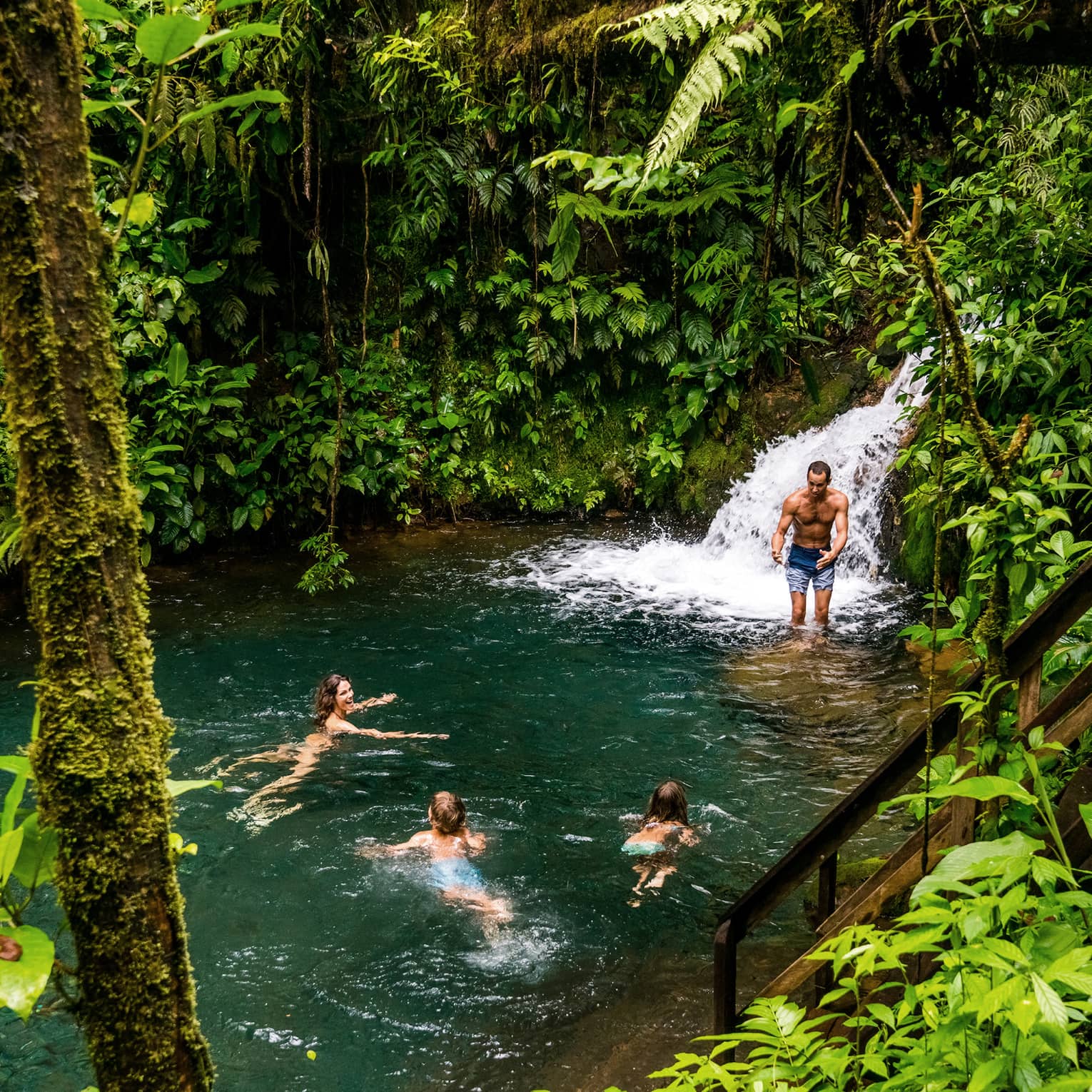 Two adults and two kids frolic at the foot of a waterfall surrounded by lush jungle greenery