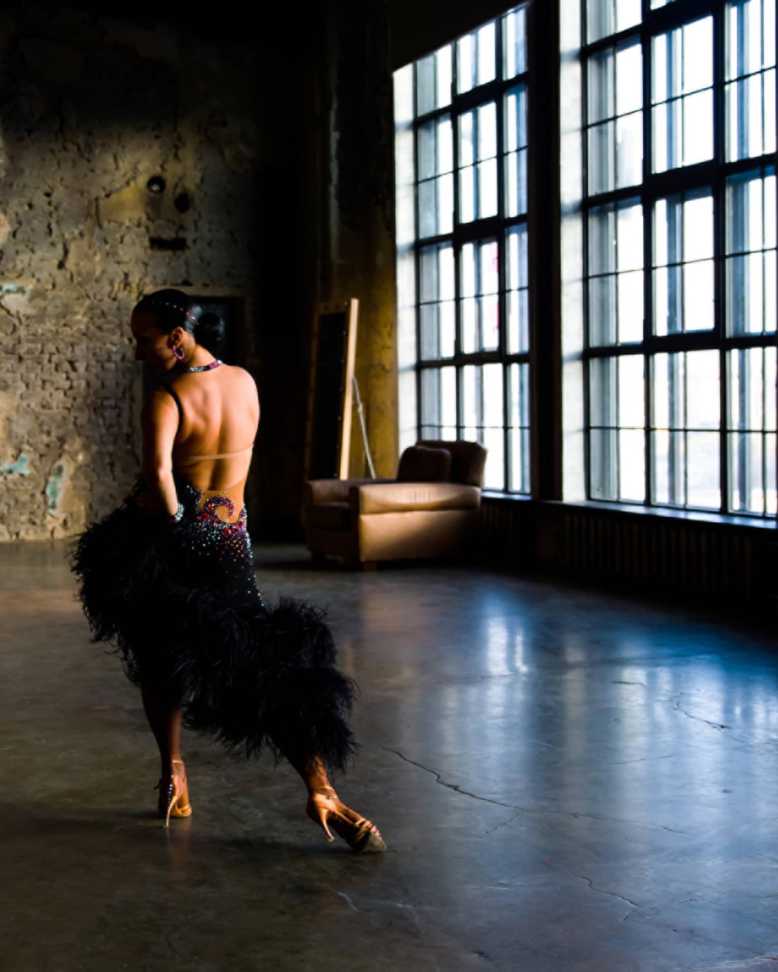 In an unlit room with large windows, a salsa dancer in an elaborate costume poses, hand on hip, opposite a brick wall.