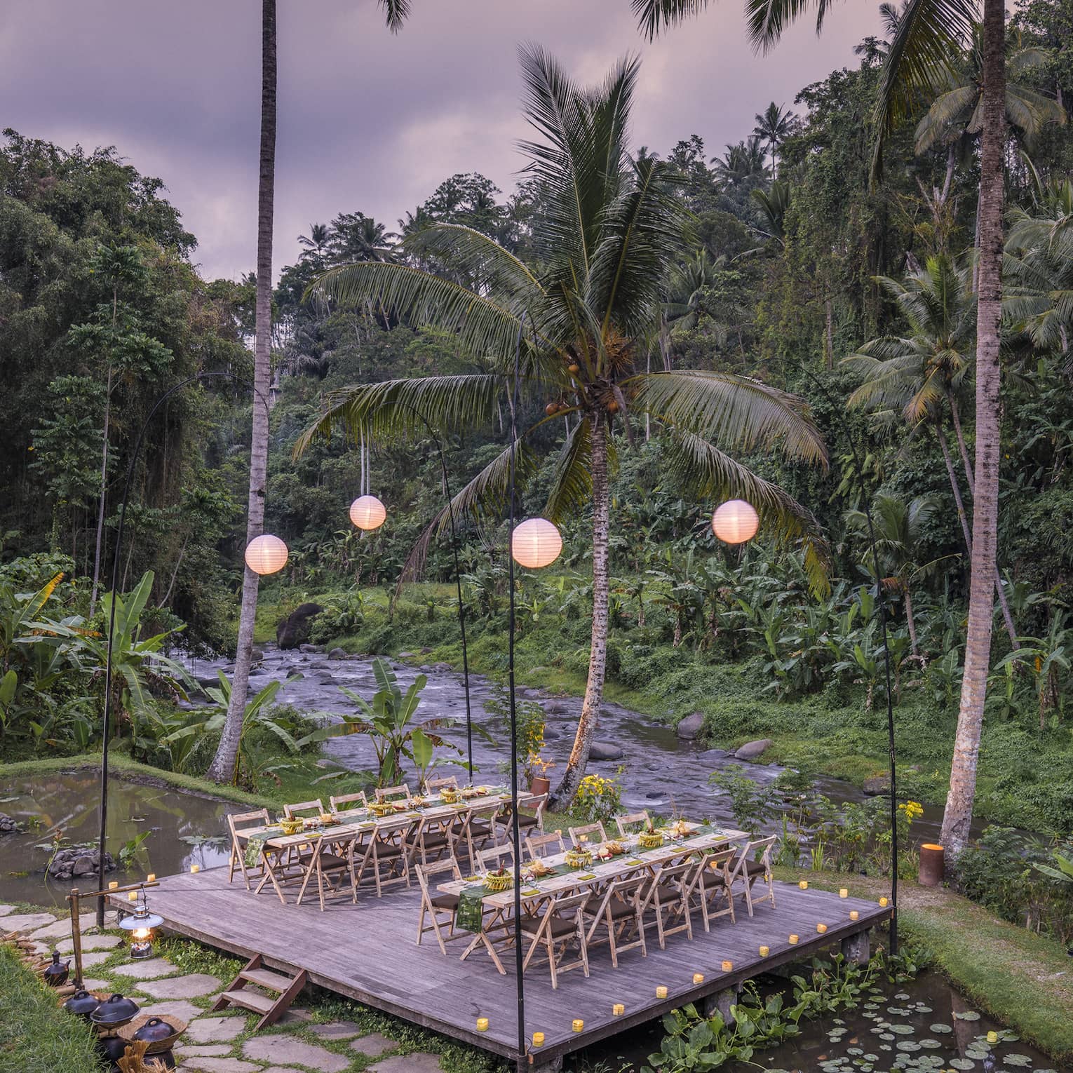 Two long tables laid out with food sit on a lantern-lit platform above a lotus pond and beside a river and dense forest.
