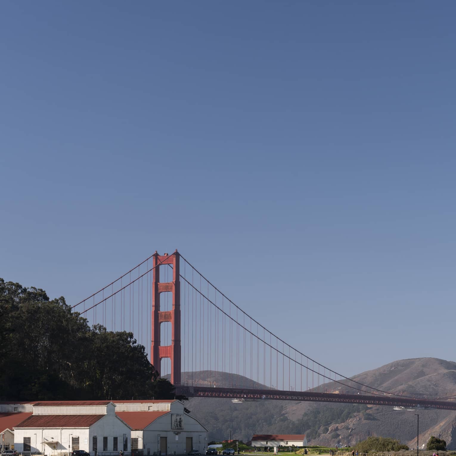 Red-roofed white buildings at the far end of a green field with the Golden Gate Bridge, rolling hills and a blue sky beyond.