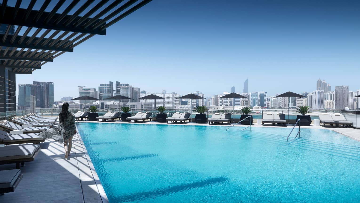 Back view of woman walking past lounge chairs on deck past blue swimming pool on sunny rooftop patio