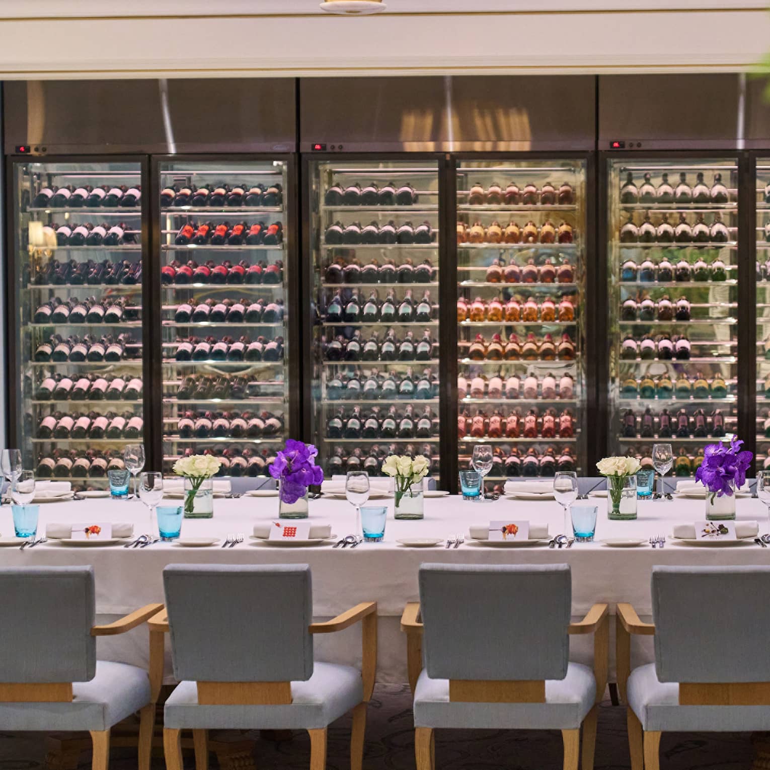 A large dining table with elegant place settings and lined by comfortable chairs before a wall of well-stocked wine fridges.
