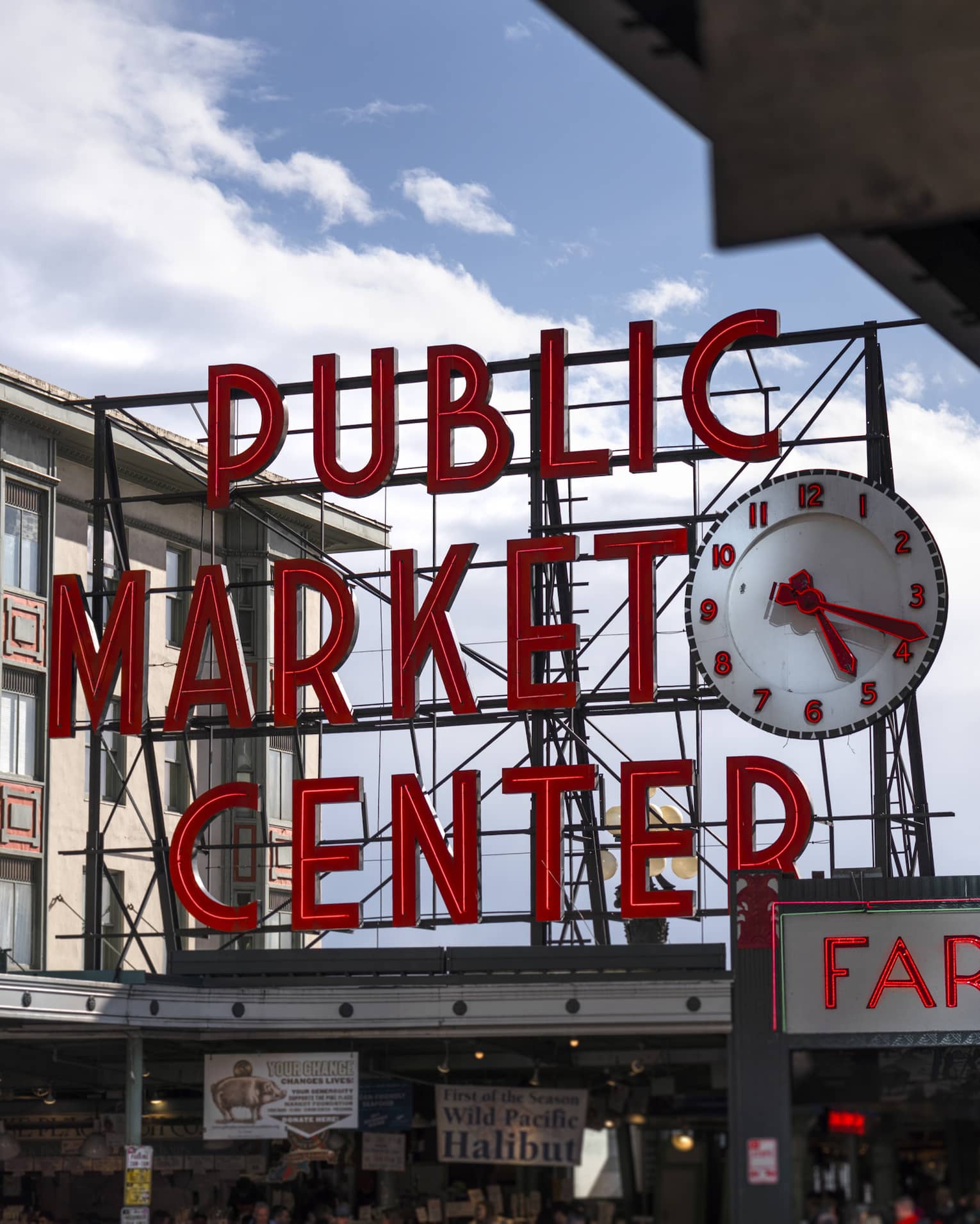 The giant Public Market Center clock and sign set in red neon letters on black scaffolding against a blue sky with clouds.