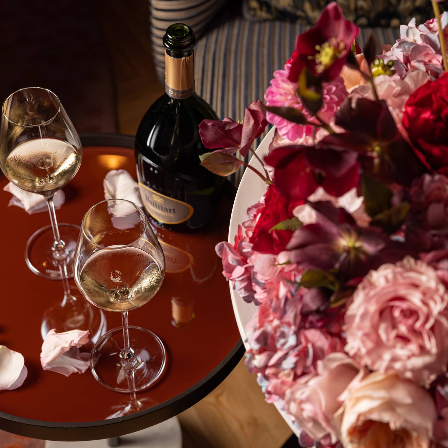 Two glasses of white wine on a brown table, surrounded by scattered rose petals and placed next to a large bouquet of flowers.