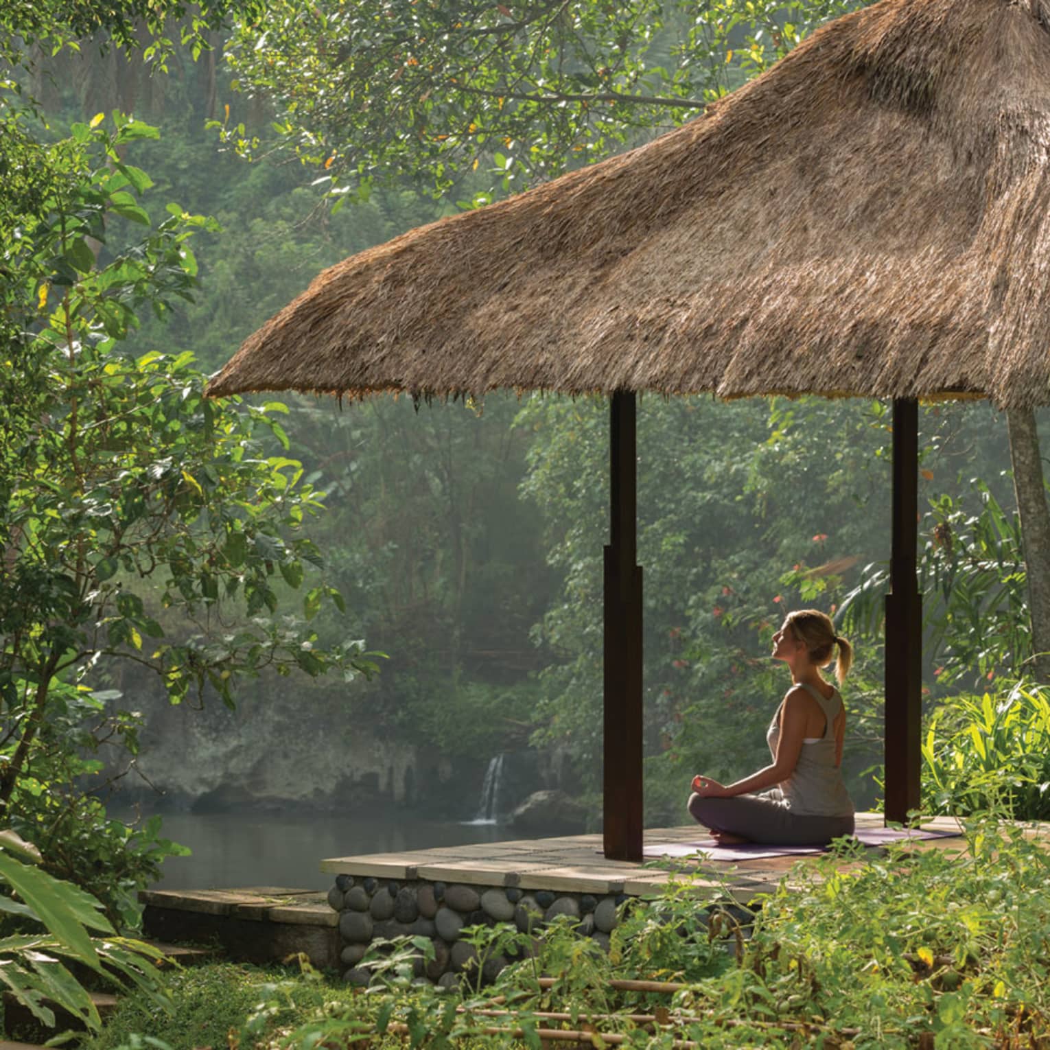 Side view of woman meditating on platform under thatched-roof gazebo, looking out at river and forest