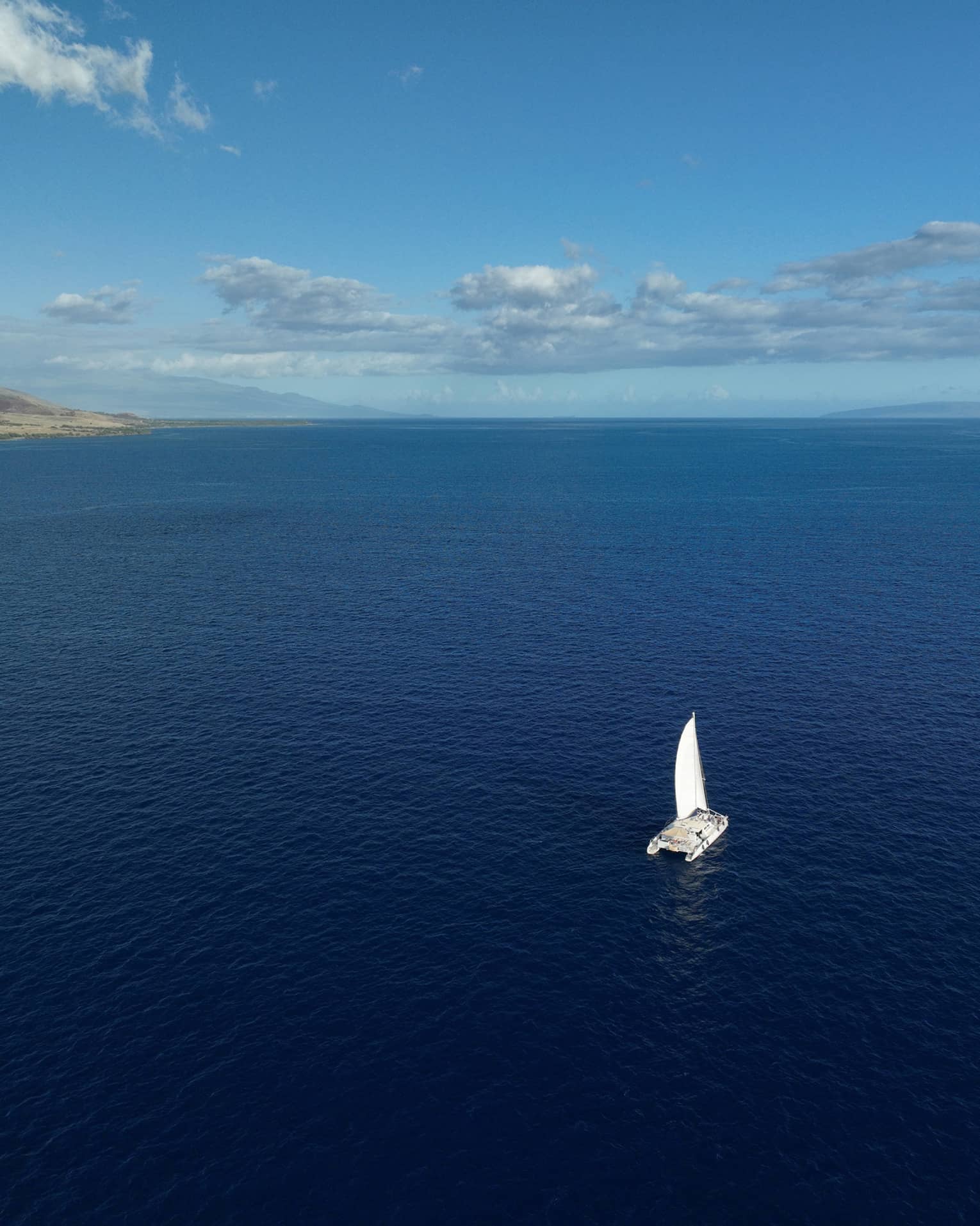 Aerial view of a catamaran as it sails through the vast, calm open waters toward the faded silhouette of distant mountains.