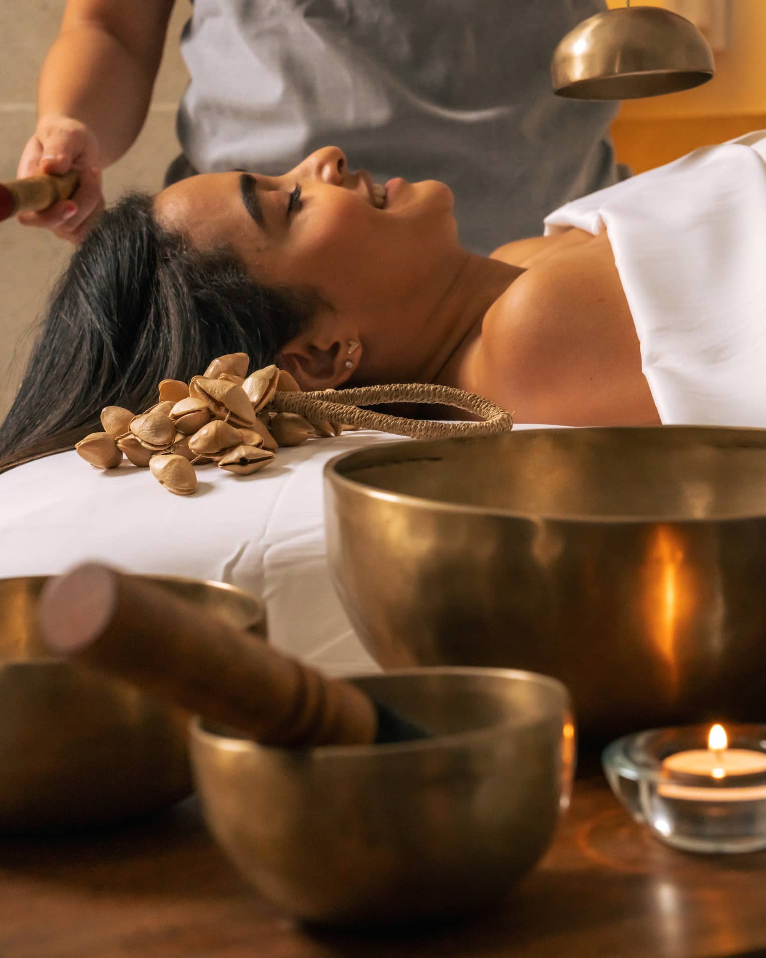 Three Tibetan sound bowls sit on a table next to a person lying beneath a white sheet while another person rings a smaller bowl over them