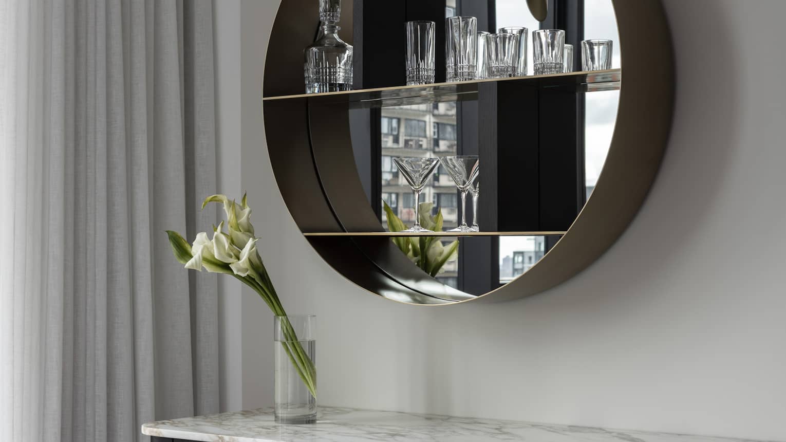 A modern vanity contains a marble countertop, round mirrored shelf displaying glassware and pink upholstered stool. A vase with white flowers sits on the vanity, complemented by soft lighting.