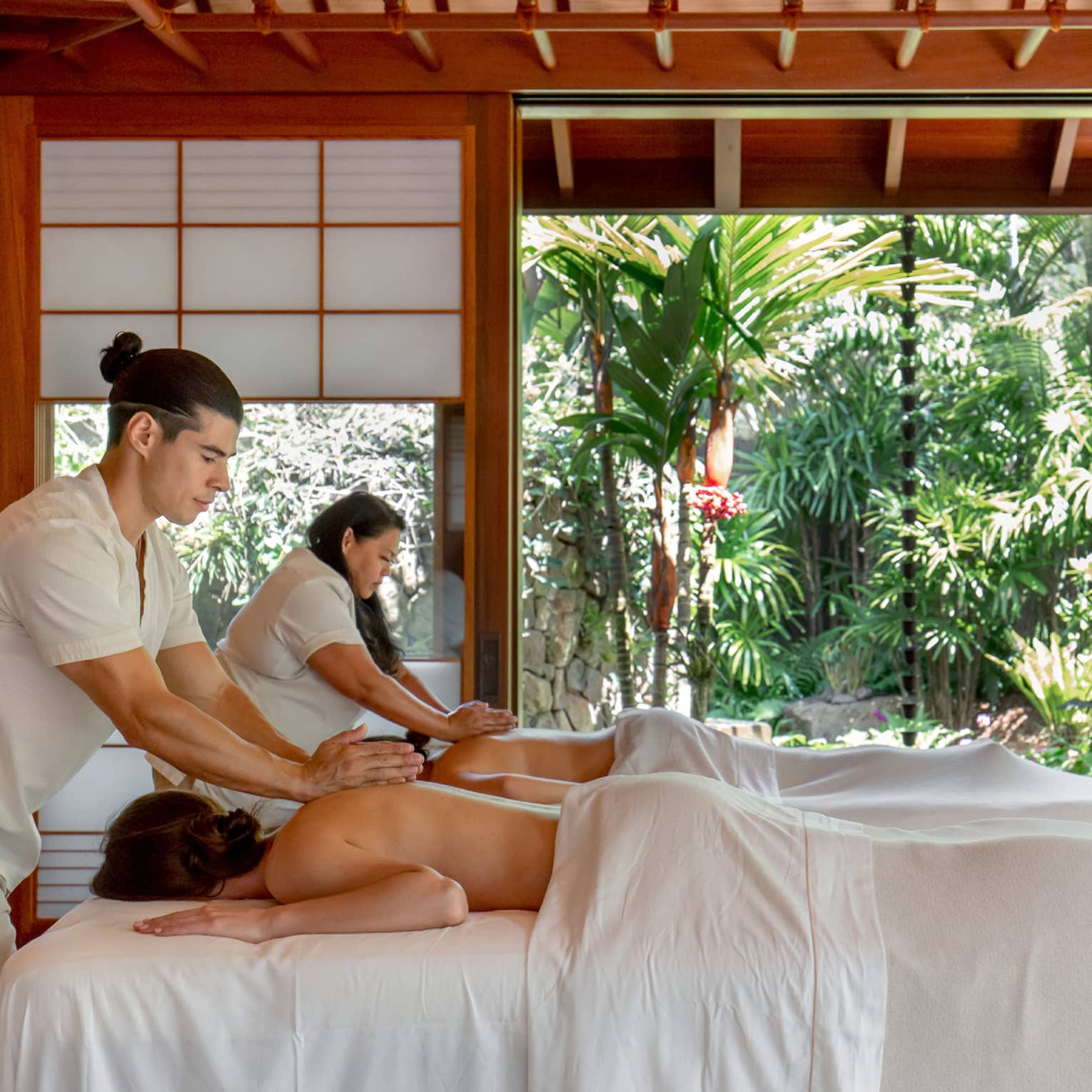 Two guests receive massages in a bright wooden hut with frosted glass windows and an entrance that opens to abundant trees.