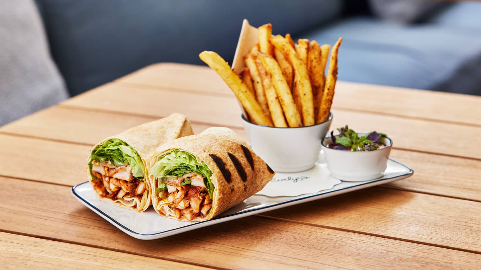 A plate with a wrap-style sandwich and a side of fries, all placed on a wood table. Upholstered seating is in the background.