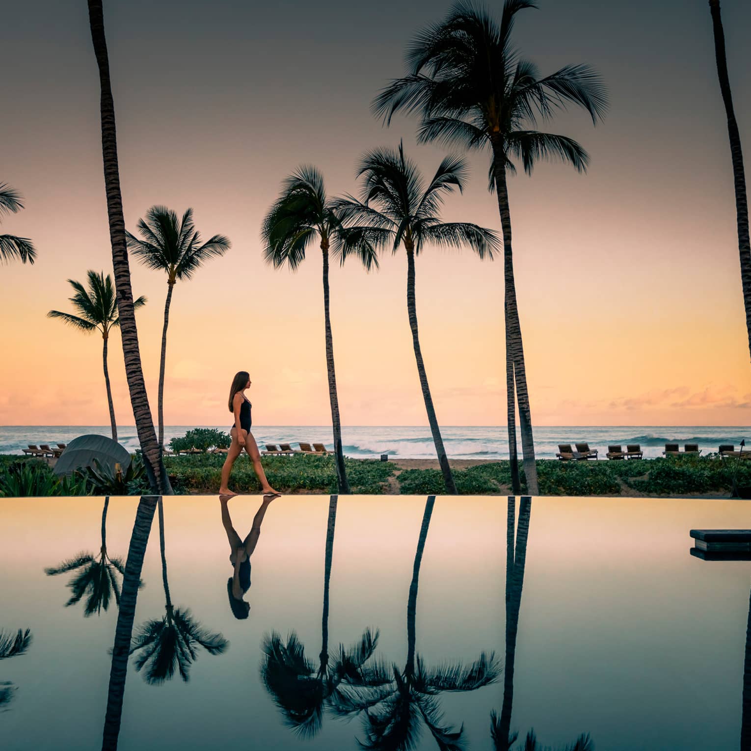 Woman in swimsuit walks by Palm Grove Pool at sunset, palm trees reflected on surface