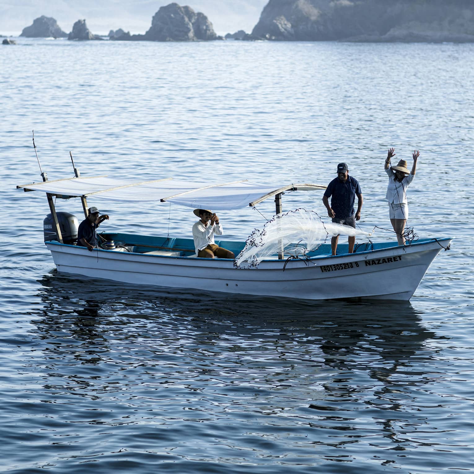 Side view of a boat with two people at the bow casting a net into the water, two others seated under a canopy near the stern.