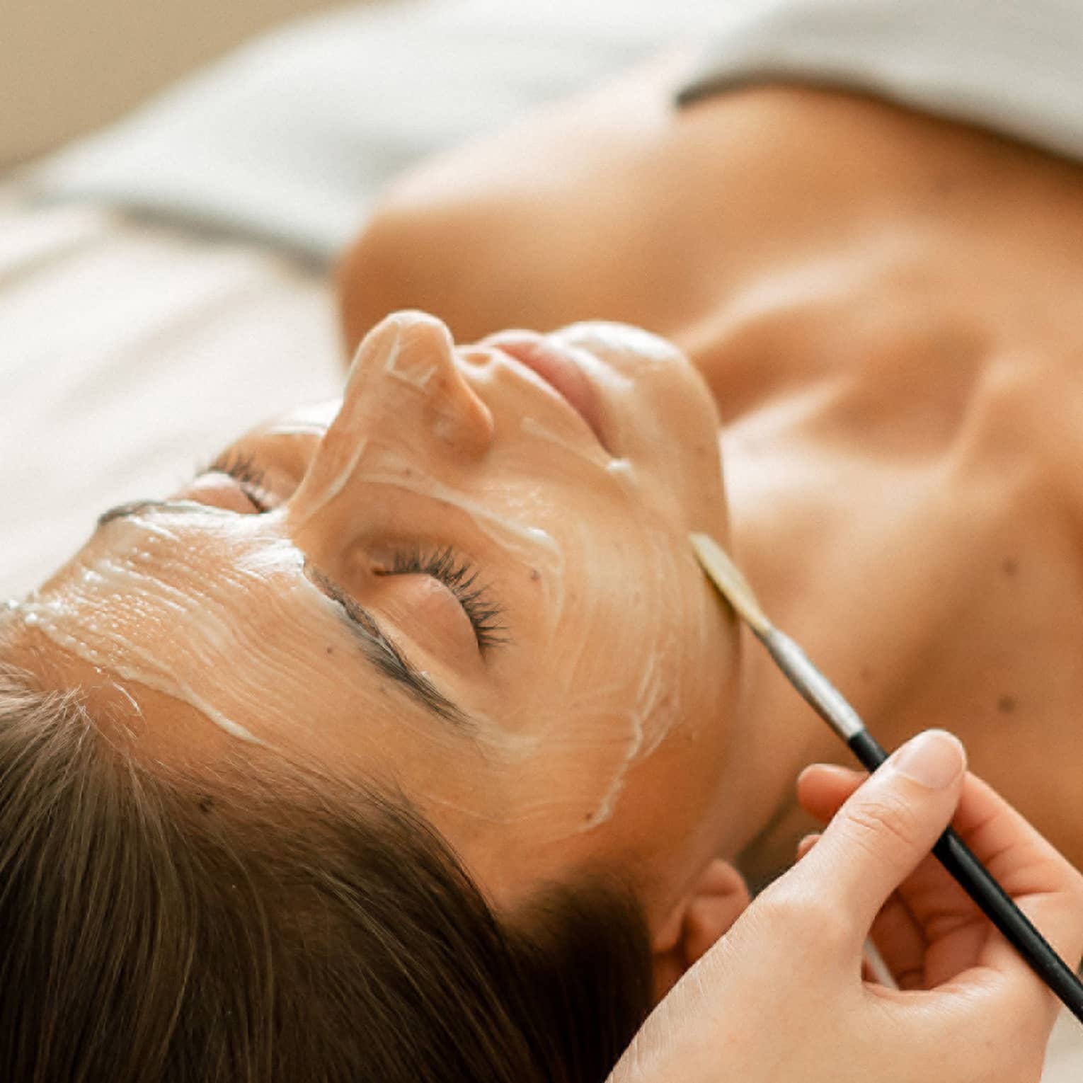 A woman lying on a spa table with eyes closed while a face mask is applied with a small brush