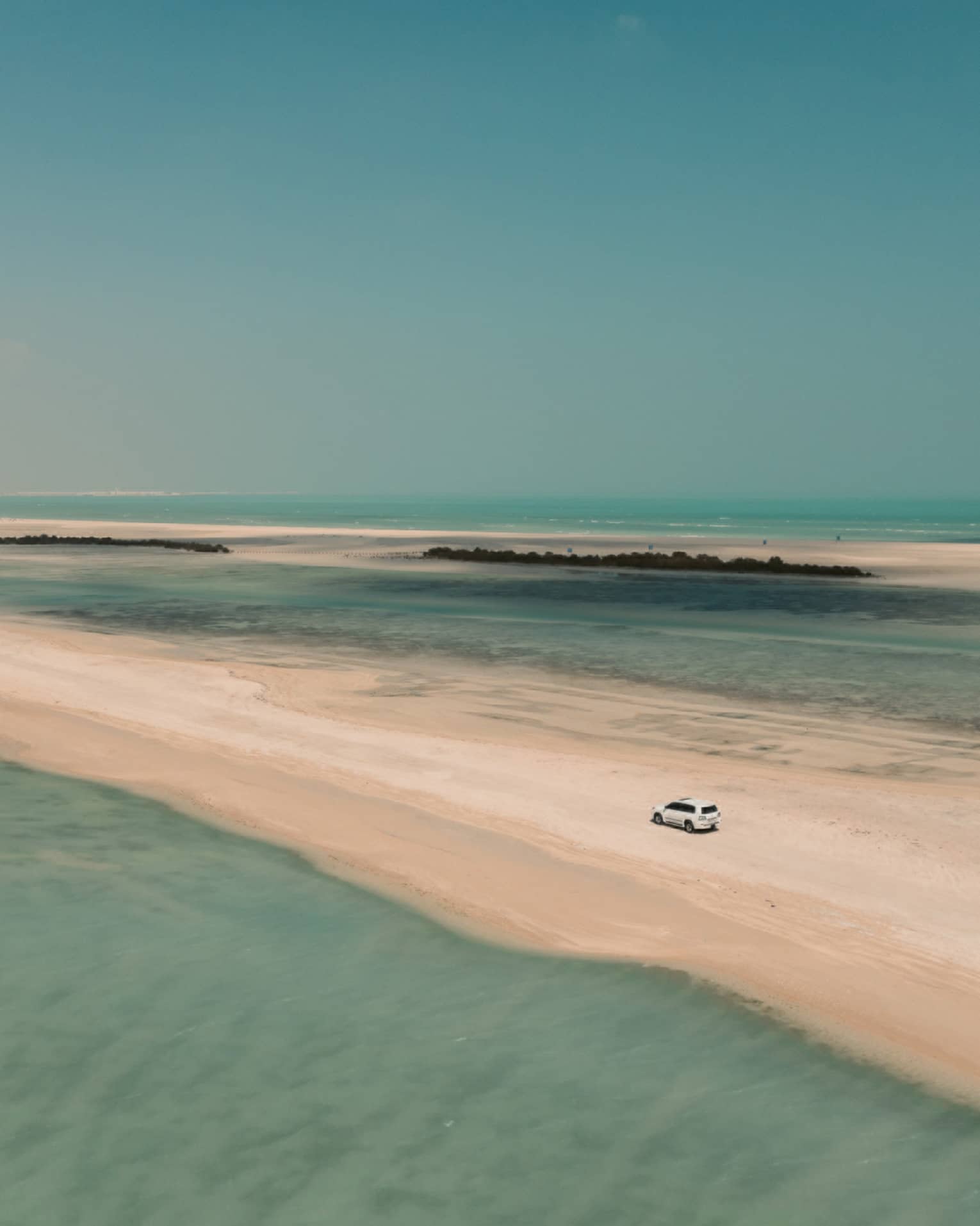 Aerial view of a 4x4 car traversing a sandbar surrounded by shallow turquoise water on either side and a cloudless sky above.