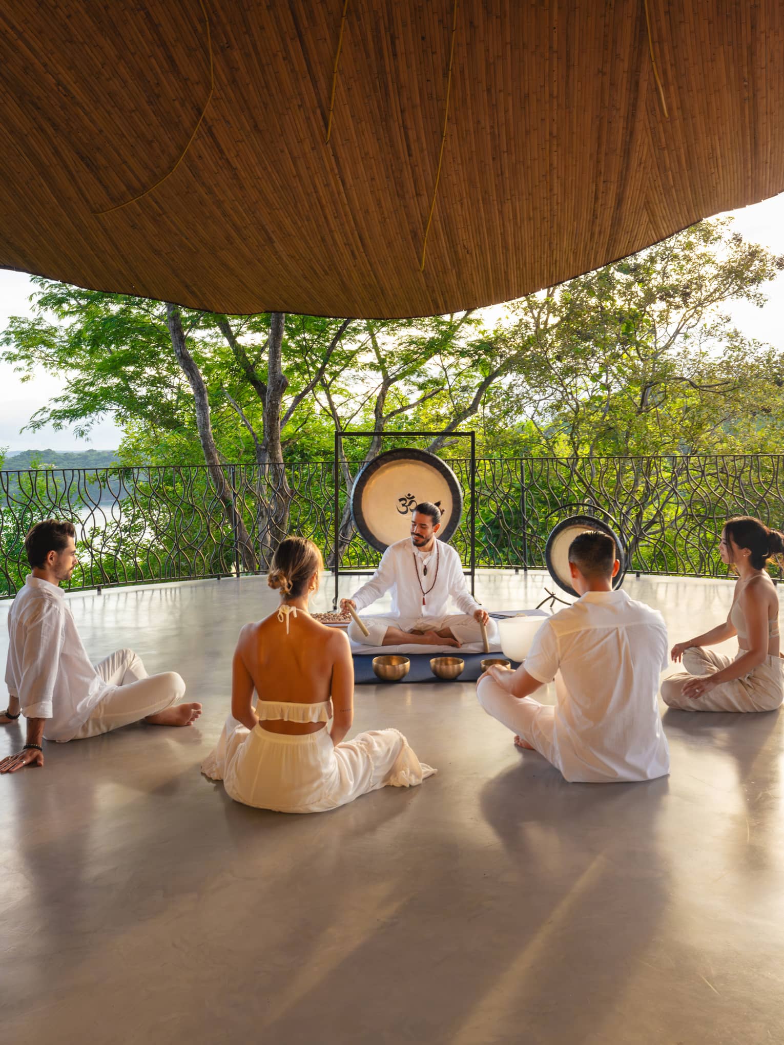 Four guests sit in a semi-circle in front of a wellness instructor who is seated in front of a gong