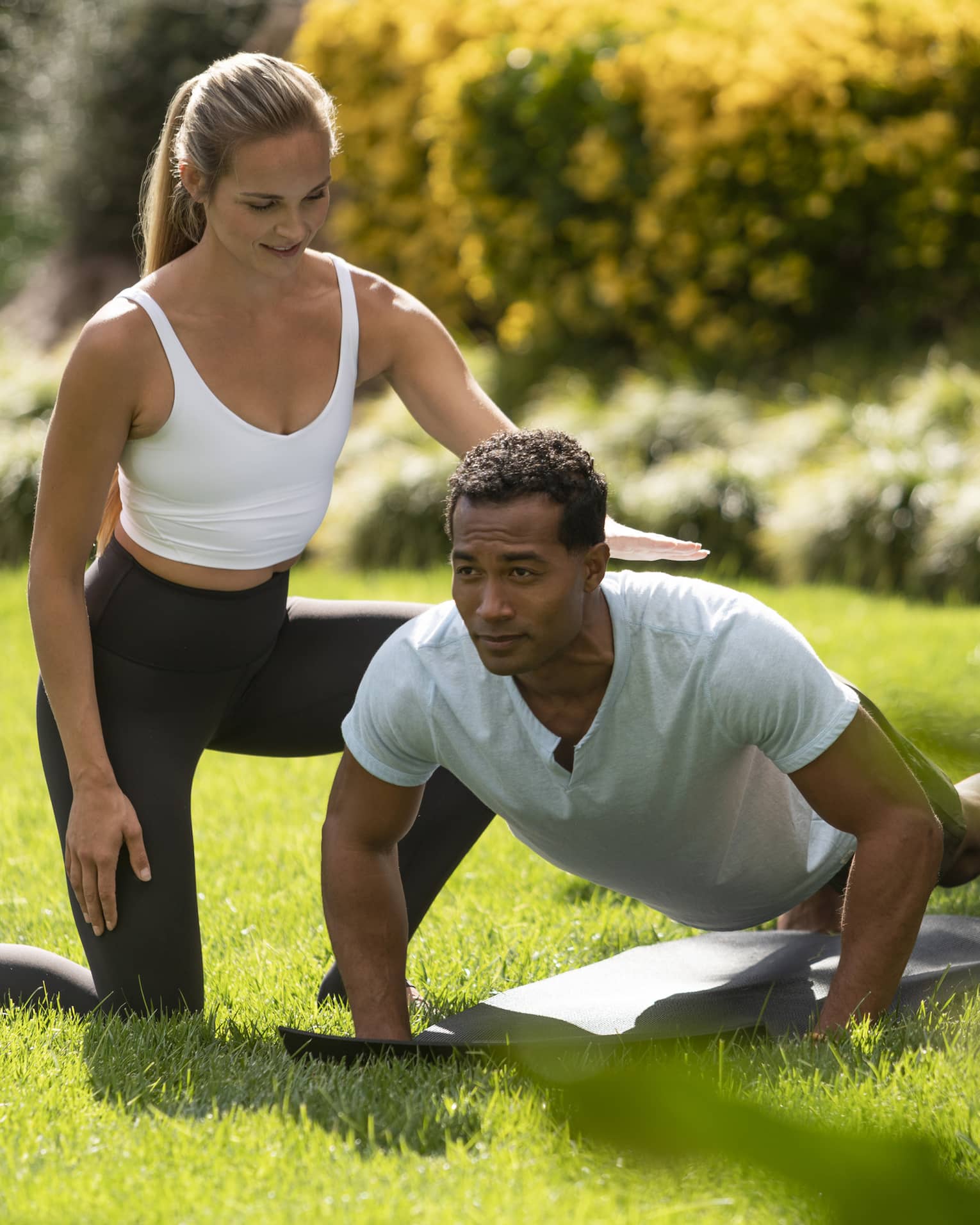 A yoga instructor helping a man get into a yoga position outside on very green grass.