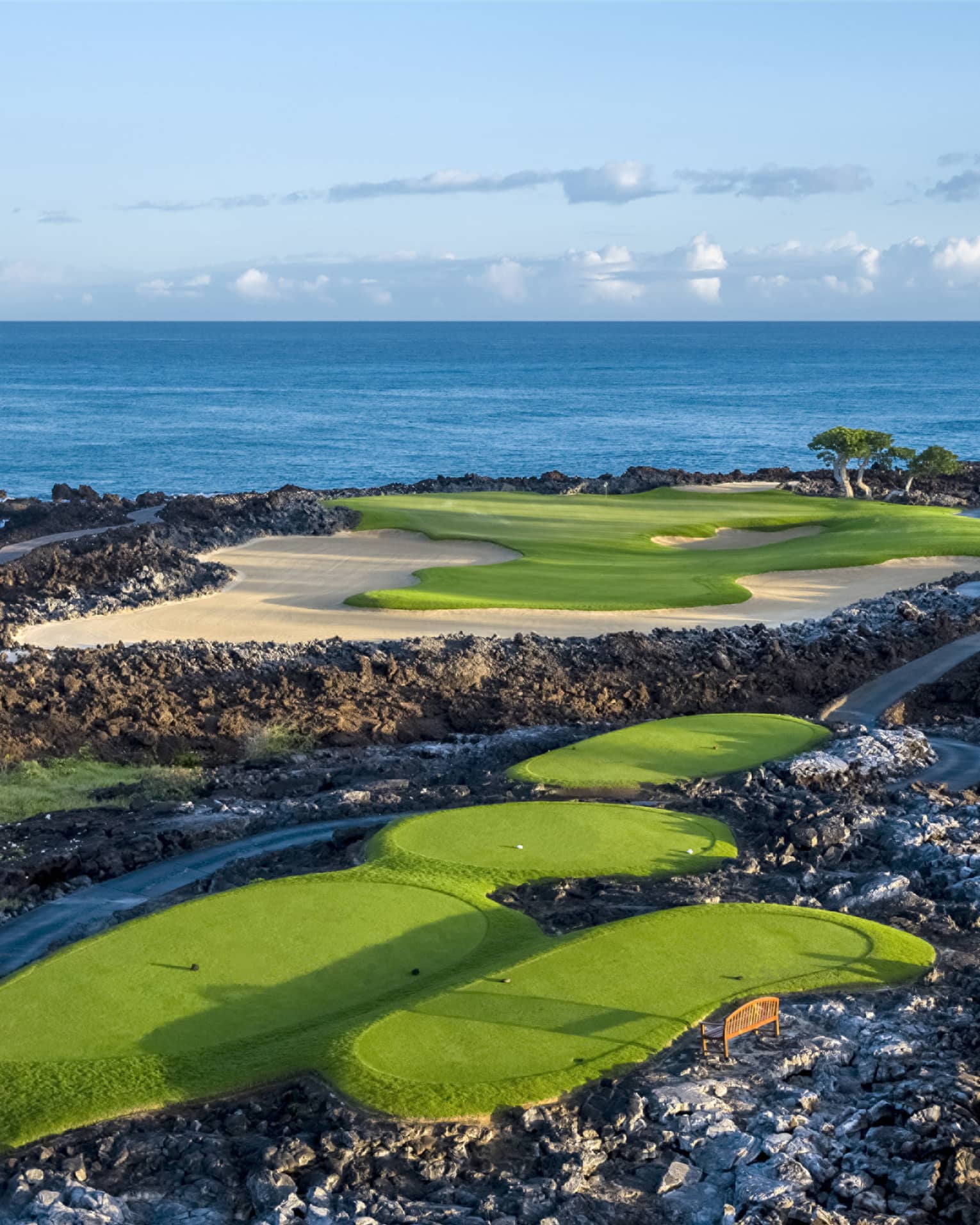 An impressive golf course overlooking the ocean, fairways and sand traps amid black lava formations, blue skies overhead.