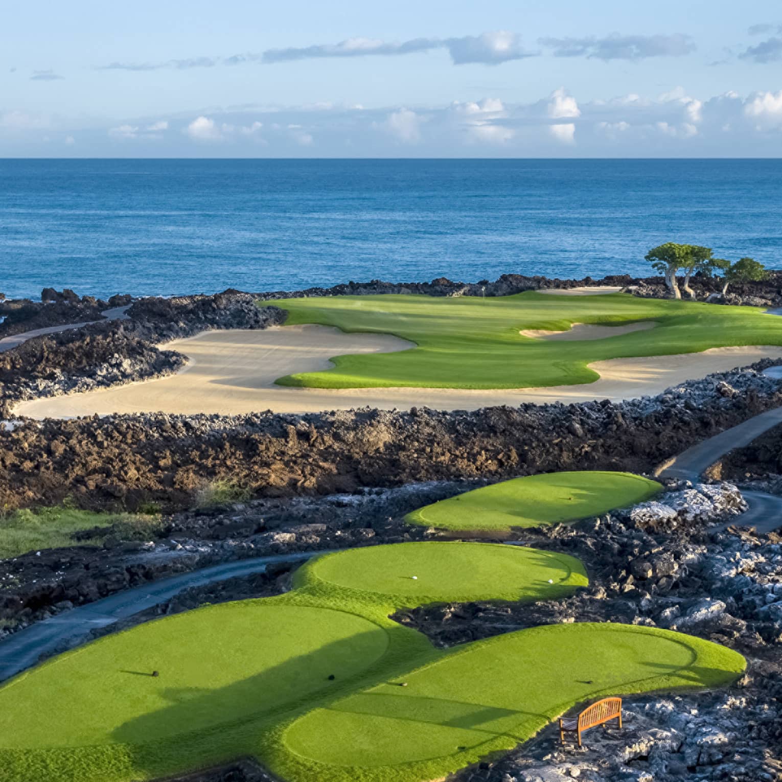An impressive golf course overlooking the ocean, fairways and sand traps amid black lava formations, blue skies overhead.