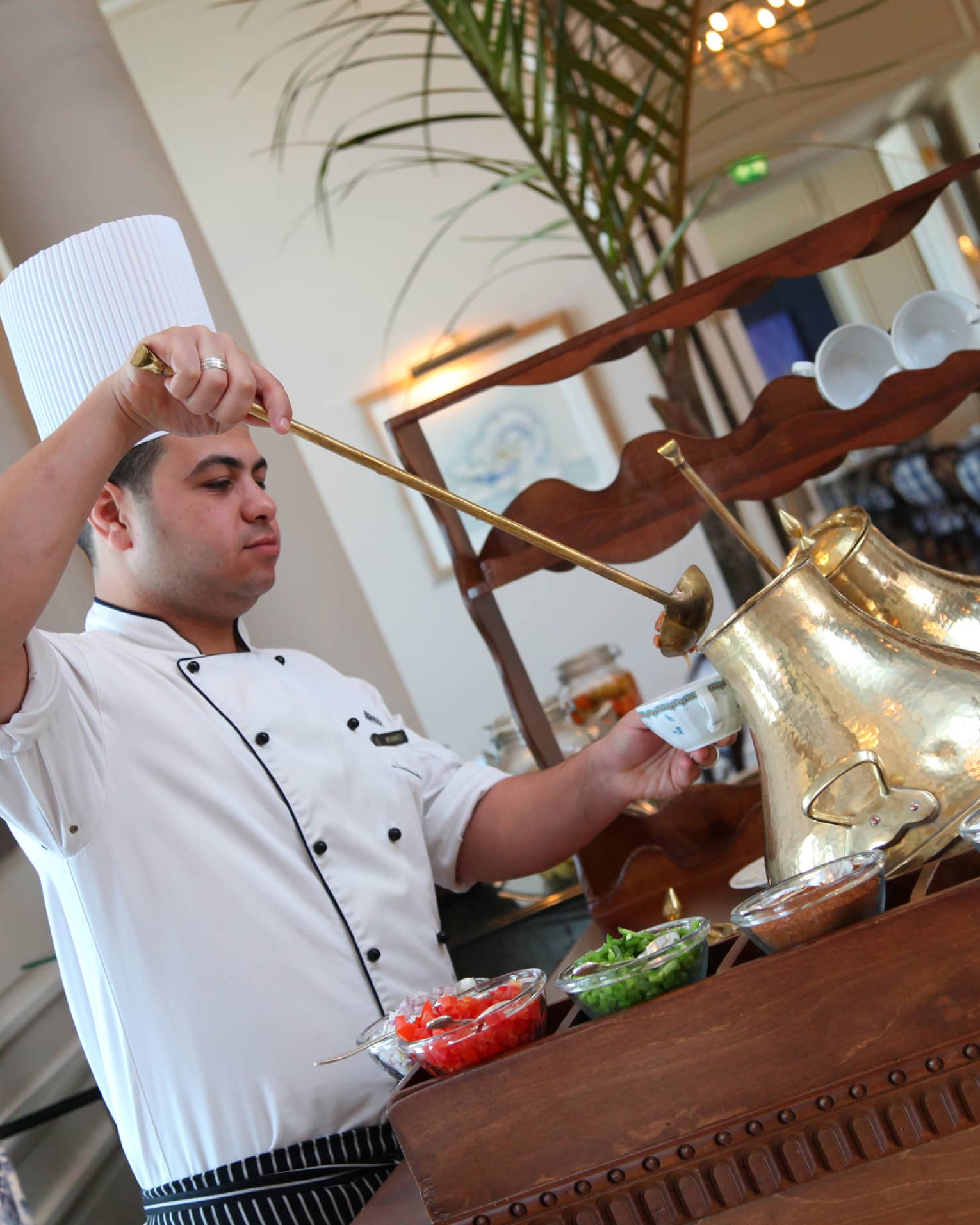 Chef in white uniform, tall hat holds long ladle, spoons liquid out of tall brass jug