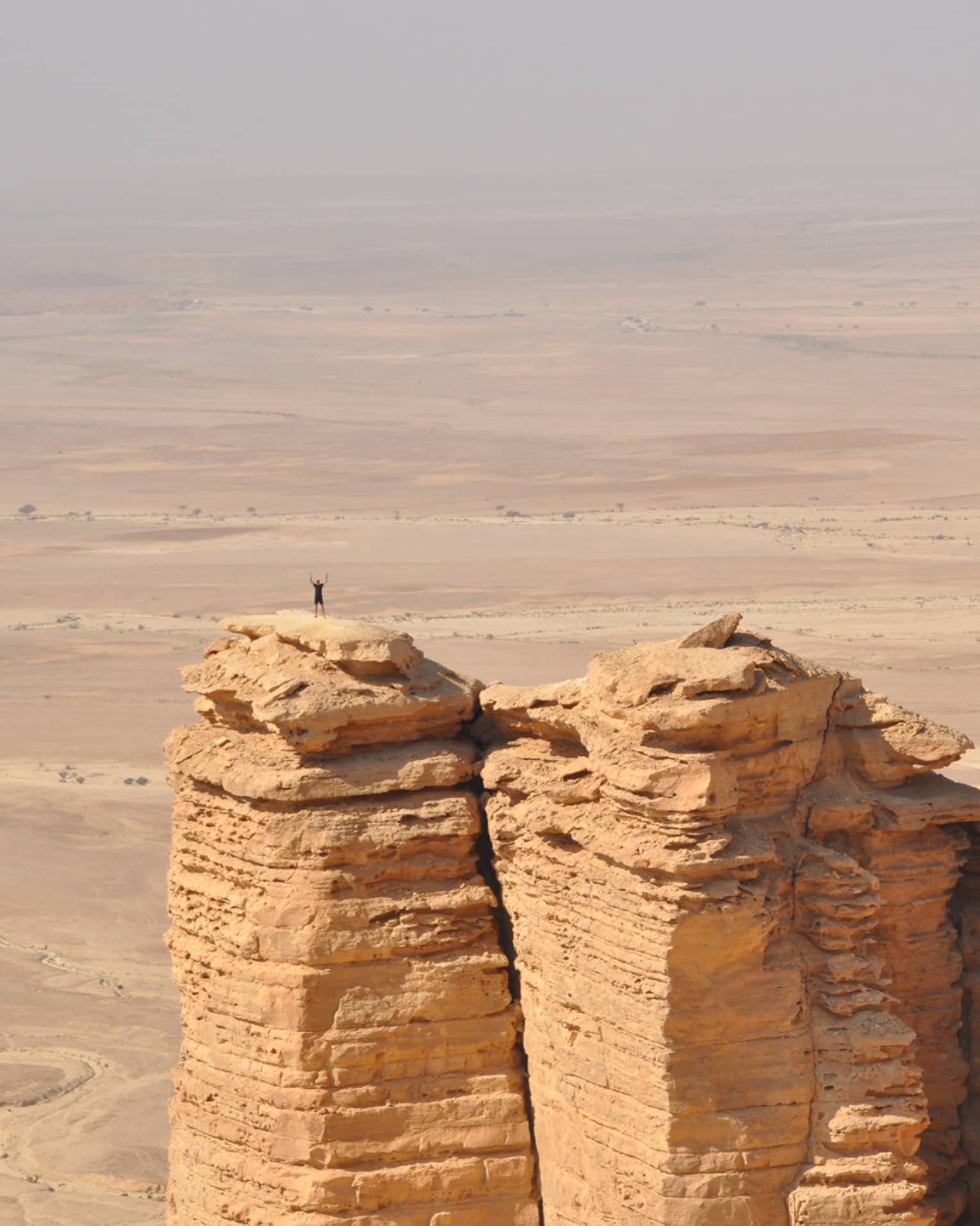 An aerial view of a giant golden cliff surrounded by a dry desert and a small silhouette of a person standing on top of it.