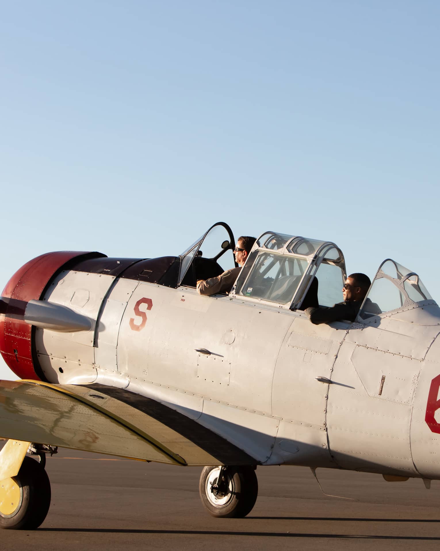 Two people sit in the cockpit of a vintage U.S. Navy aircraft on a runway, preparing for takeoff under a clear sky.