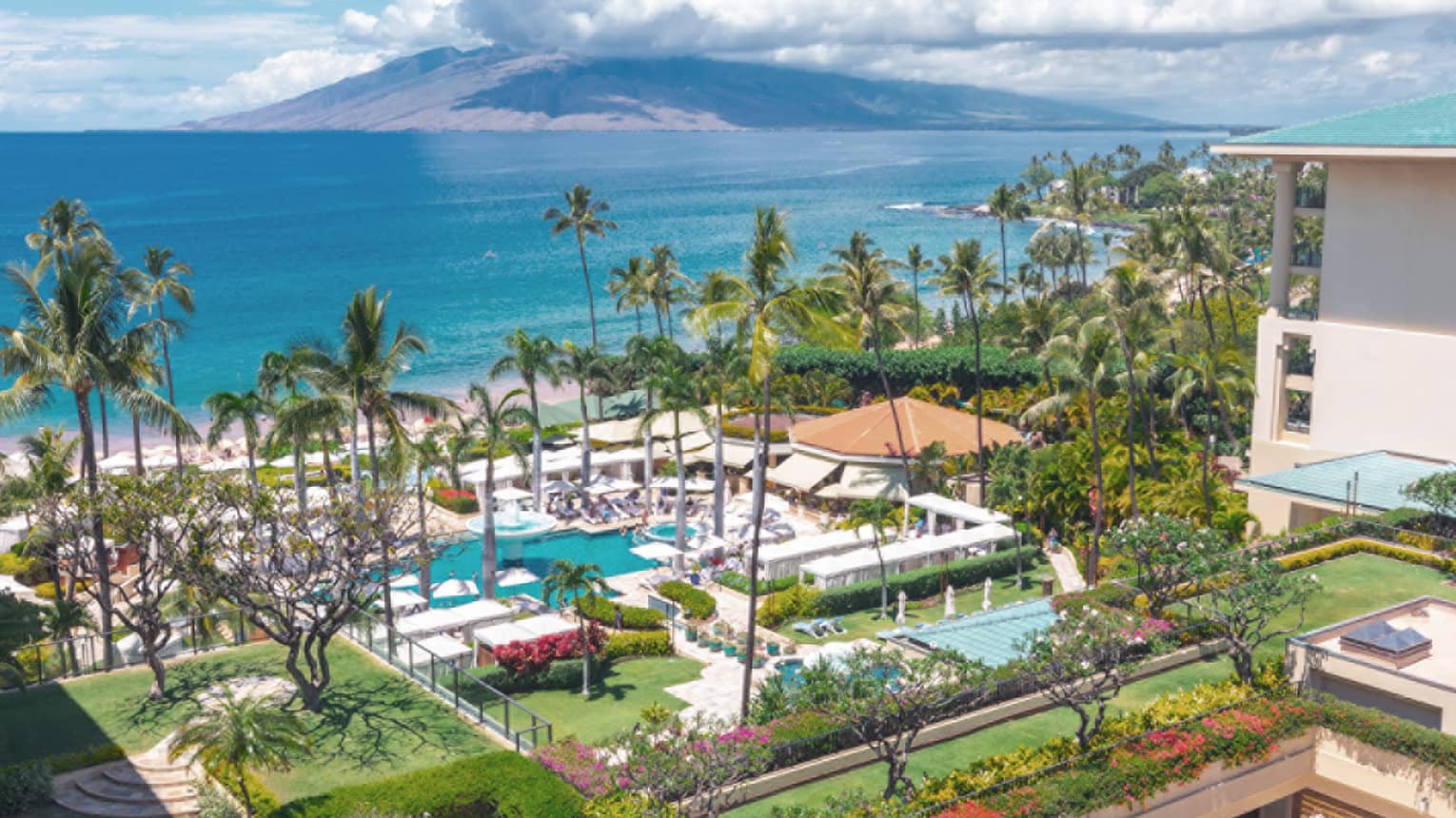 Aerial view of resort with pools, palm trees, gardens and beach along a tropical coastline with mountains in the distance