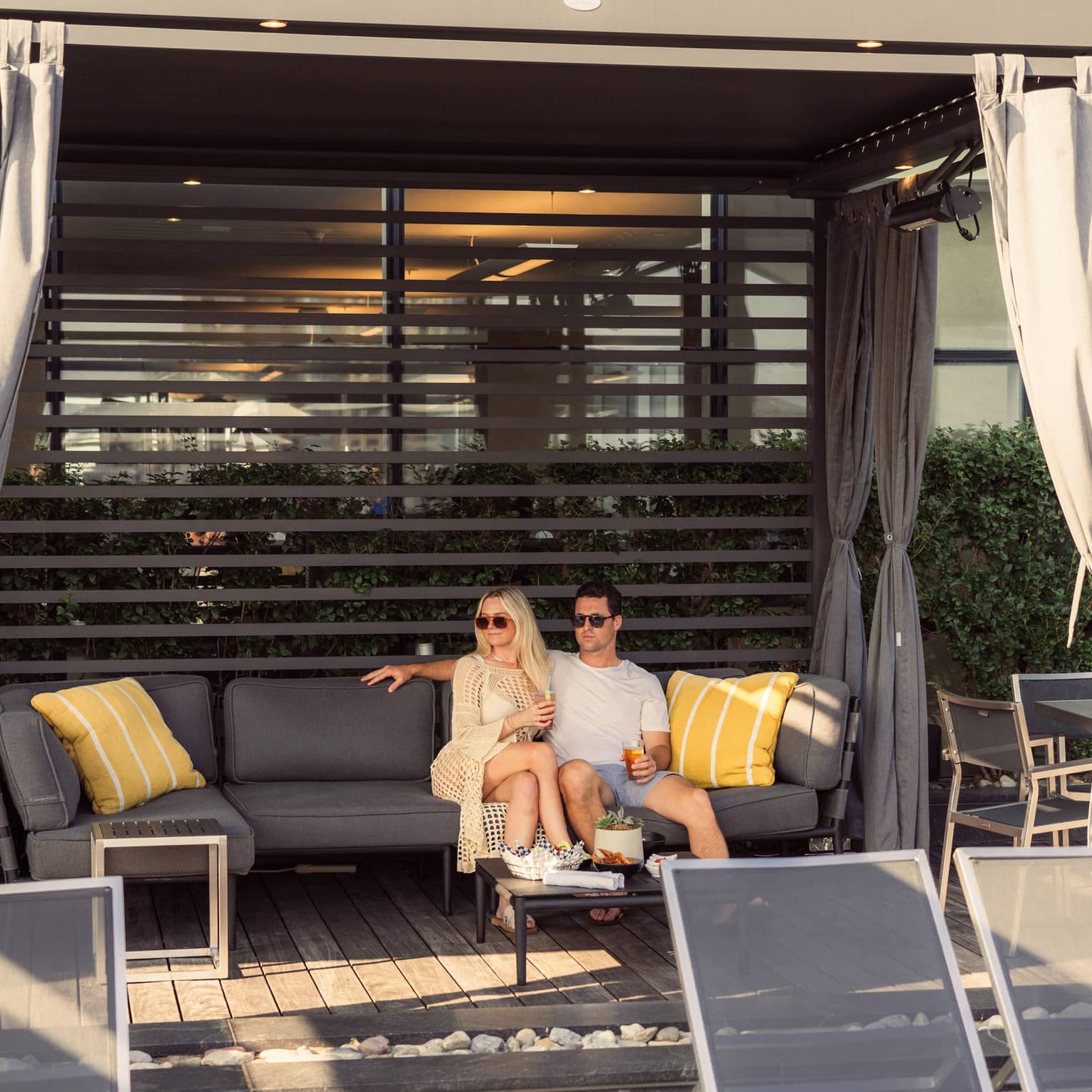 In a sun-dappled cabana, a couple relaxes together, drinks in hand, on a plush patio sofa, a table of snacks set for them.