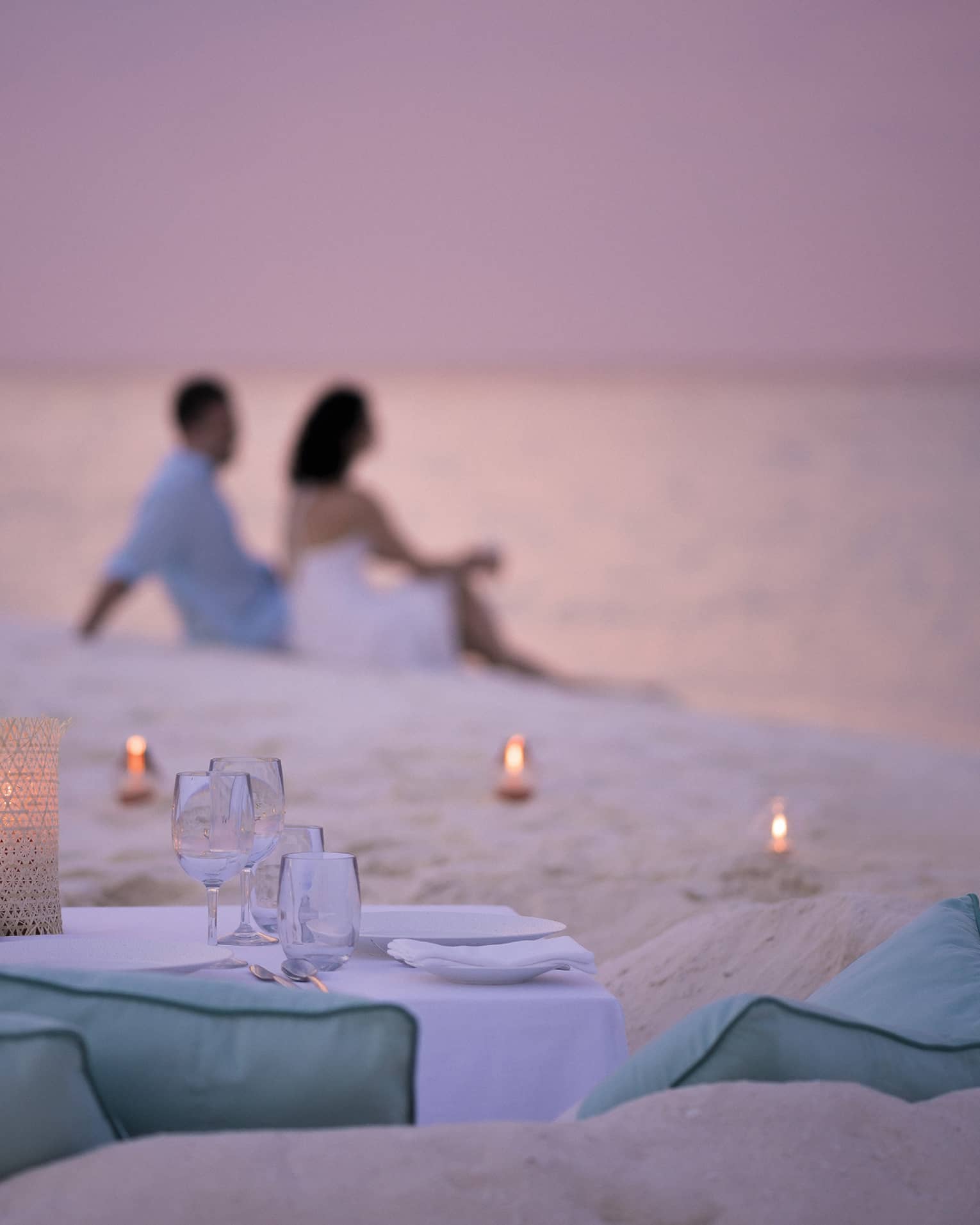 Private dining table in front of sand beach with tealight candles, couple sitting in sand