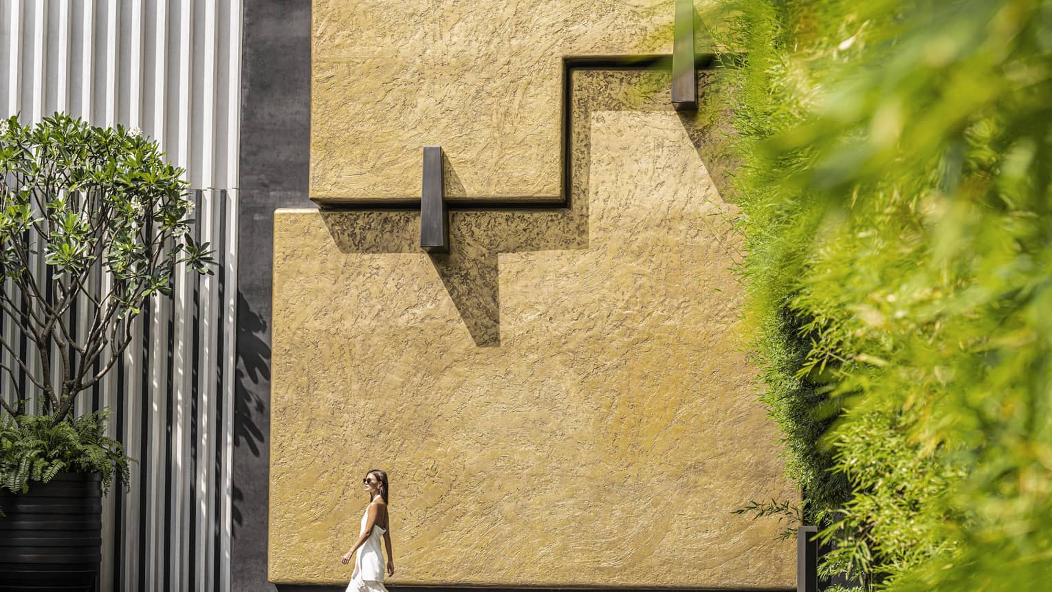 Woman walks around the exterior of the Hotel, stucco wall, oversized potted greenery