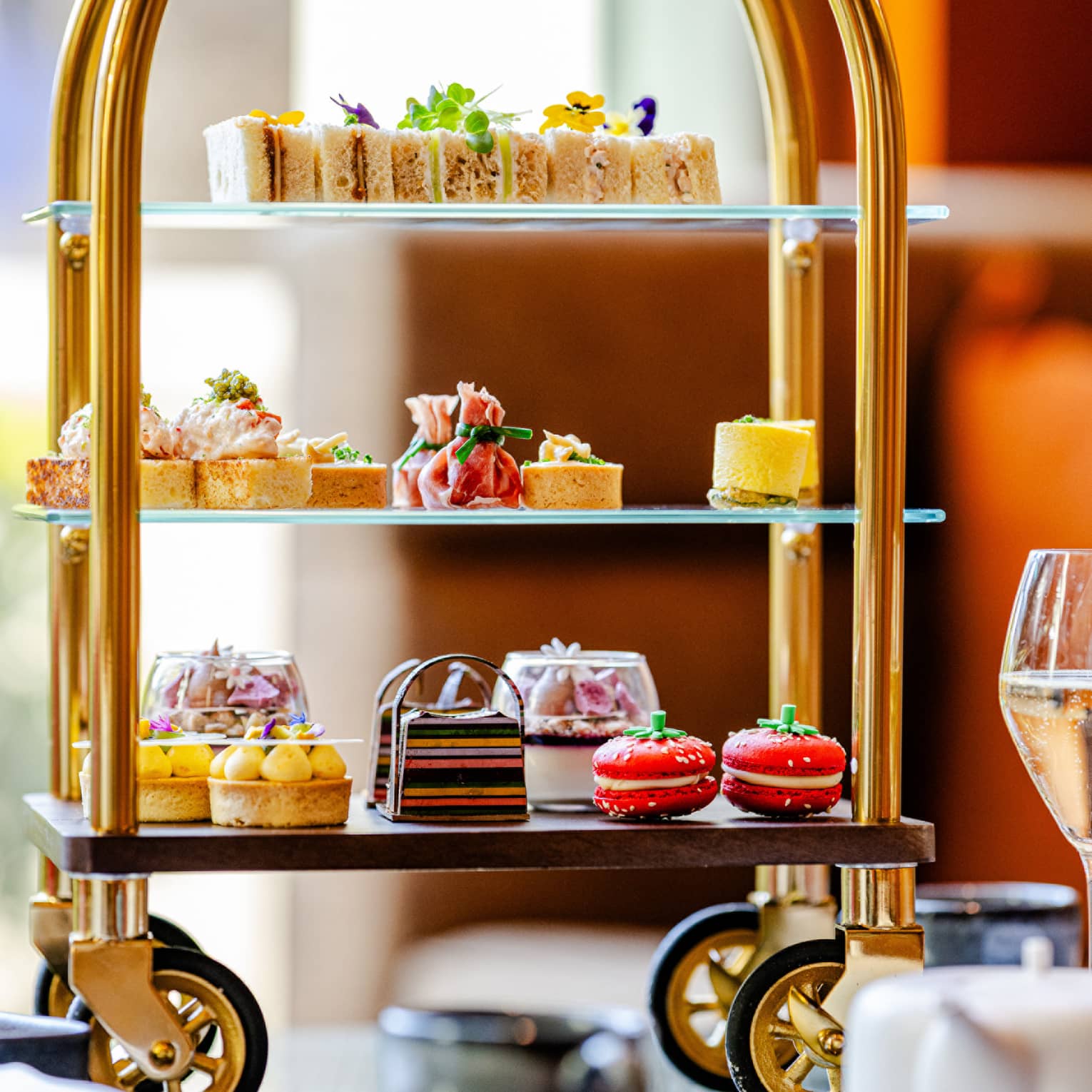 A tiered serving tray on small wheels holds an assortment of colourful small bites. It sits on a tabletop next to a stemmed glass, a teapot, and a teacup and saucer.