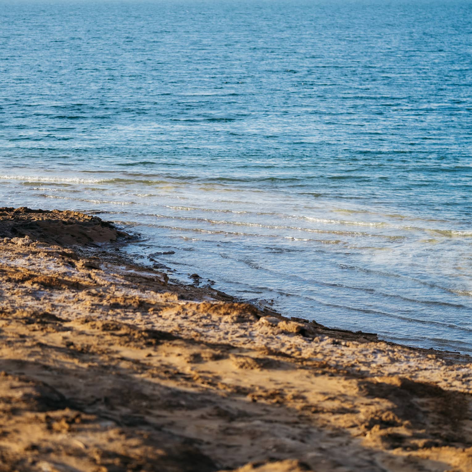 Gentle waves in sparkling azure water undulate toward an empty dark-brown, rough-sand beach.